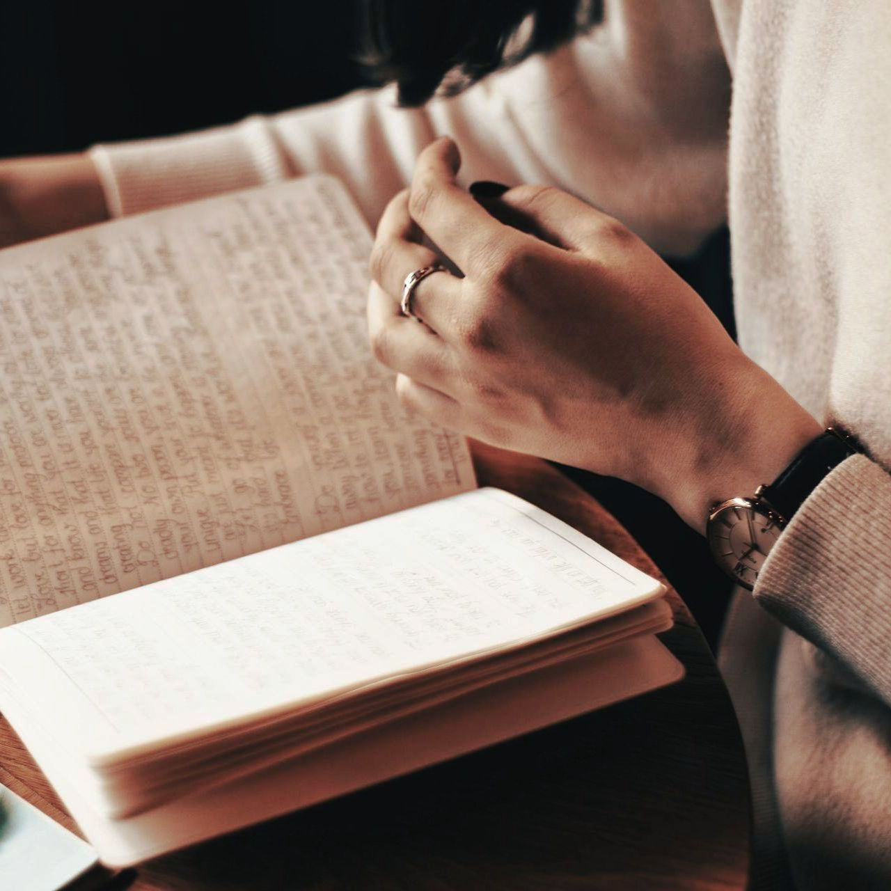 Frau liest ein Buch, hält die Seiten in den Händen, trägt einen Ring und eine Uhr und einen weißen Pullover.