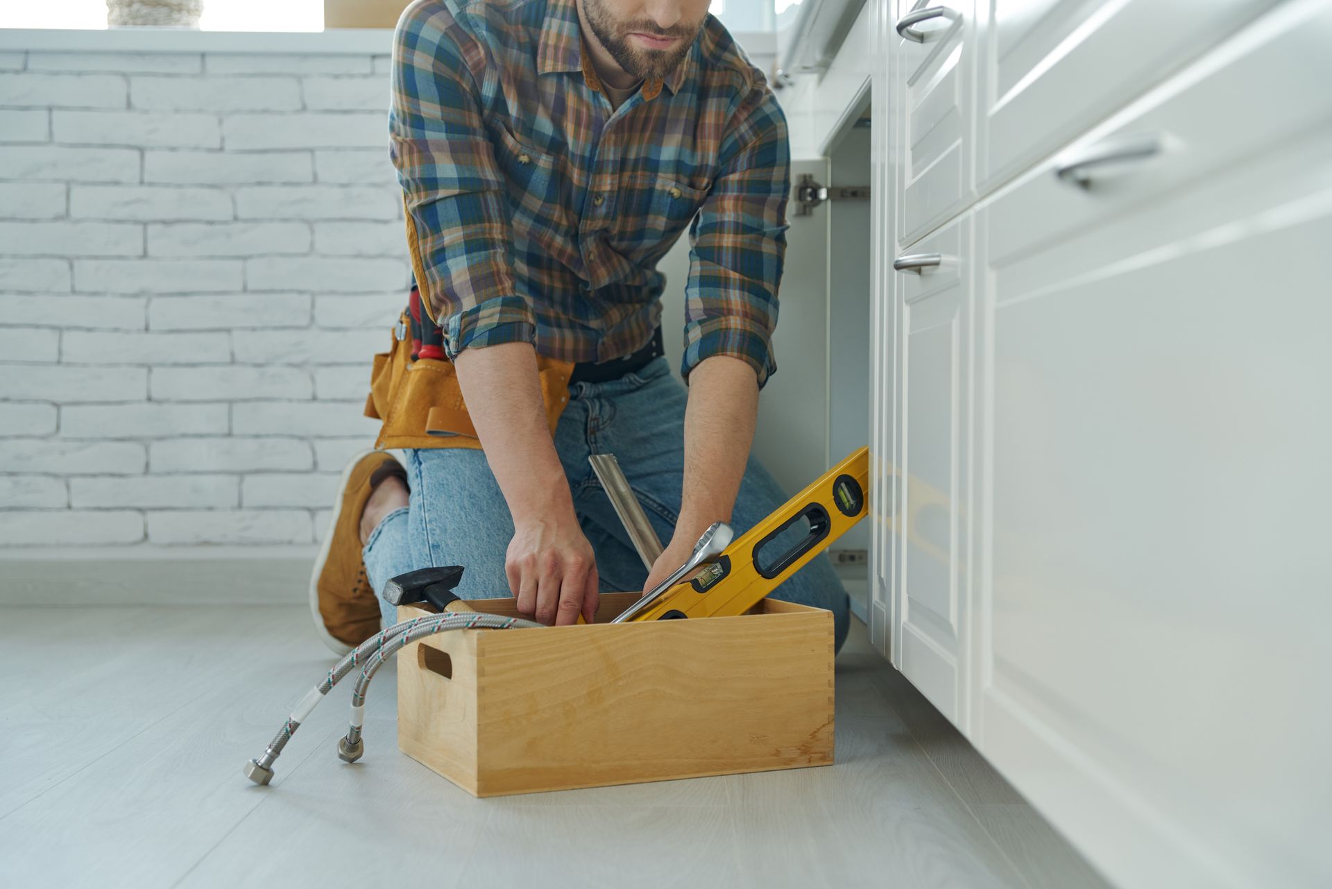 Person with tool belt kneels to retrieve tools from a wooden box under a white cabinet.