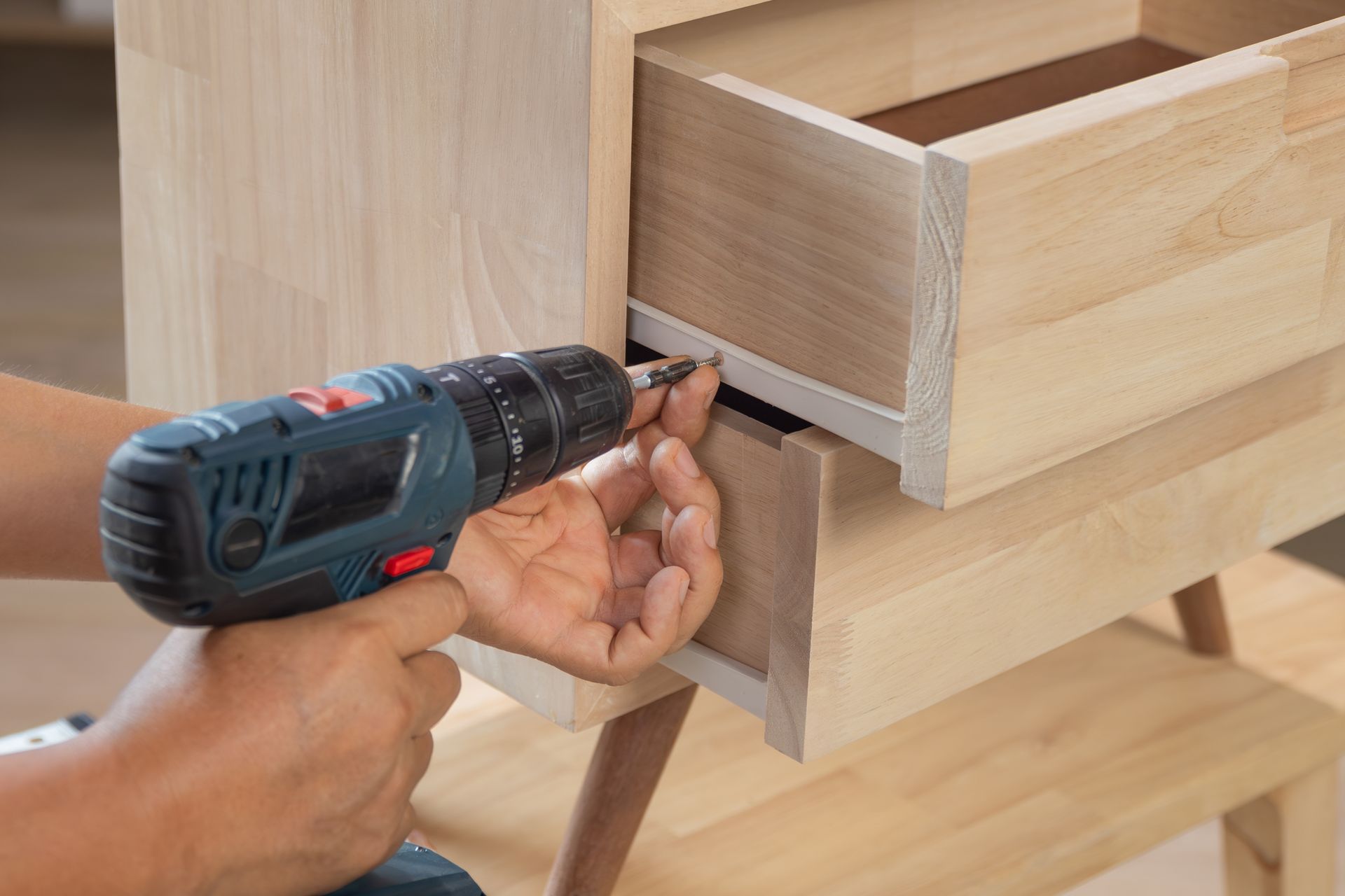 Person using a power drill to assemble a wooden drawer in a light-colored wooden cabinet.