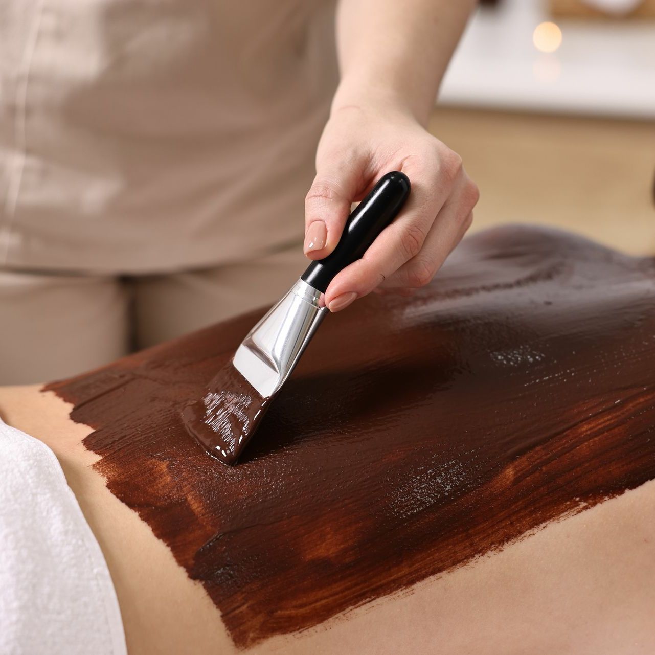 Person applying a dark brown mud mask to a person's back with a spatula.