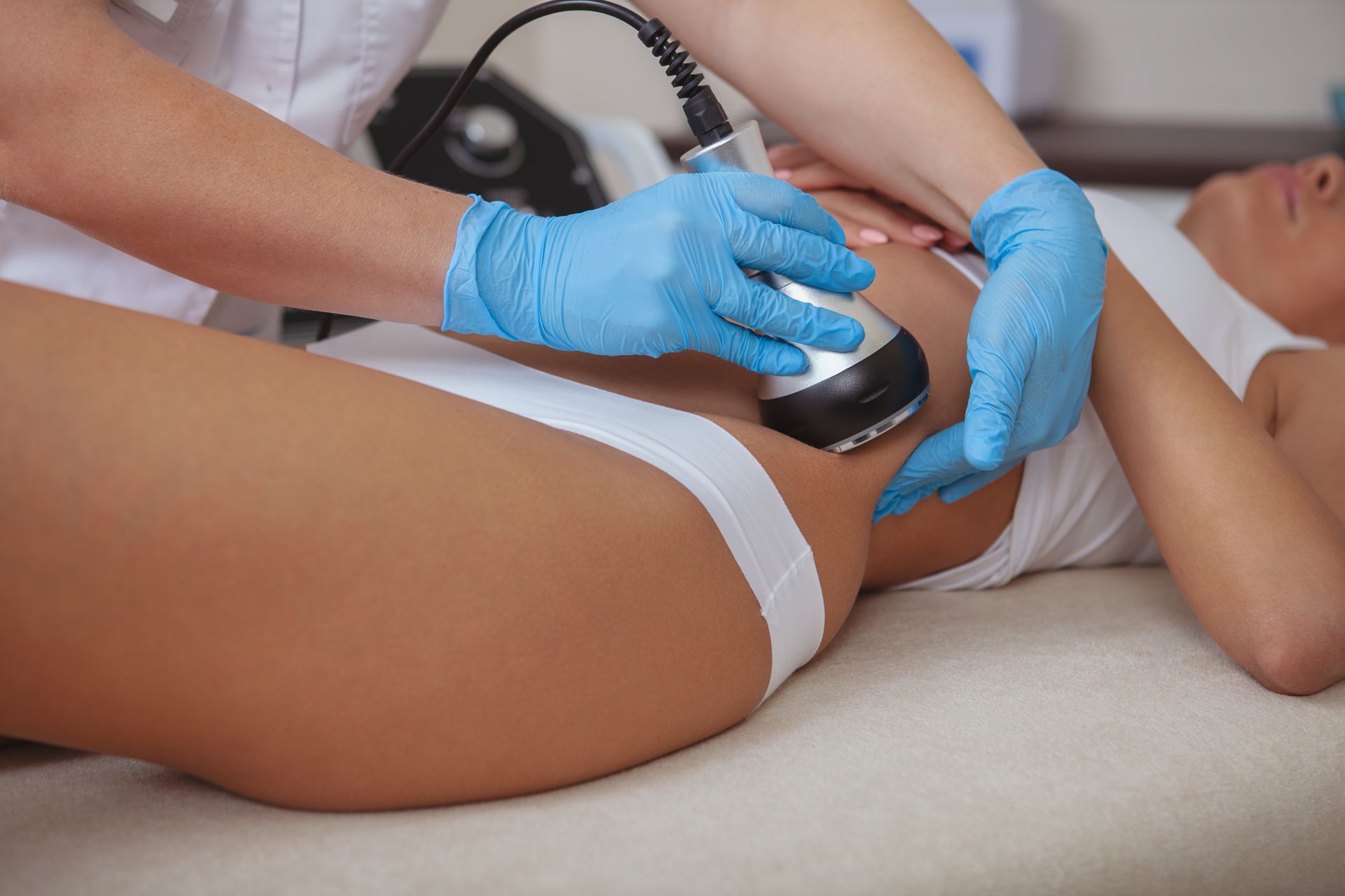 A technician using a machine on a woman's hip, possibly for body contouring. Blue gloves, white clothing.