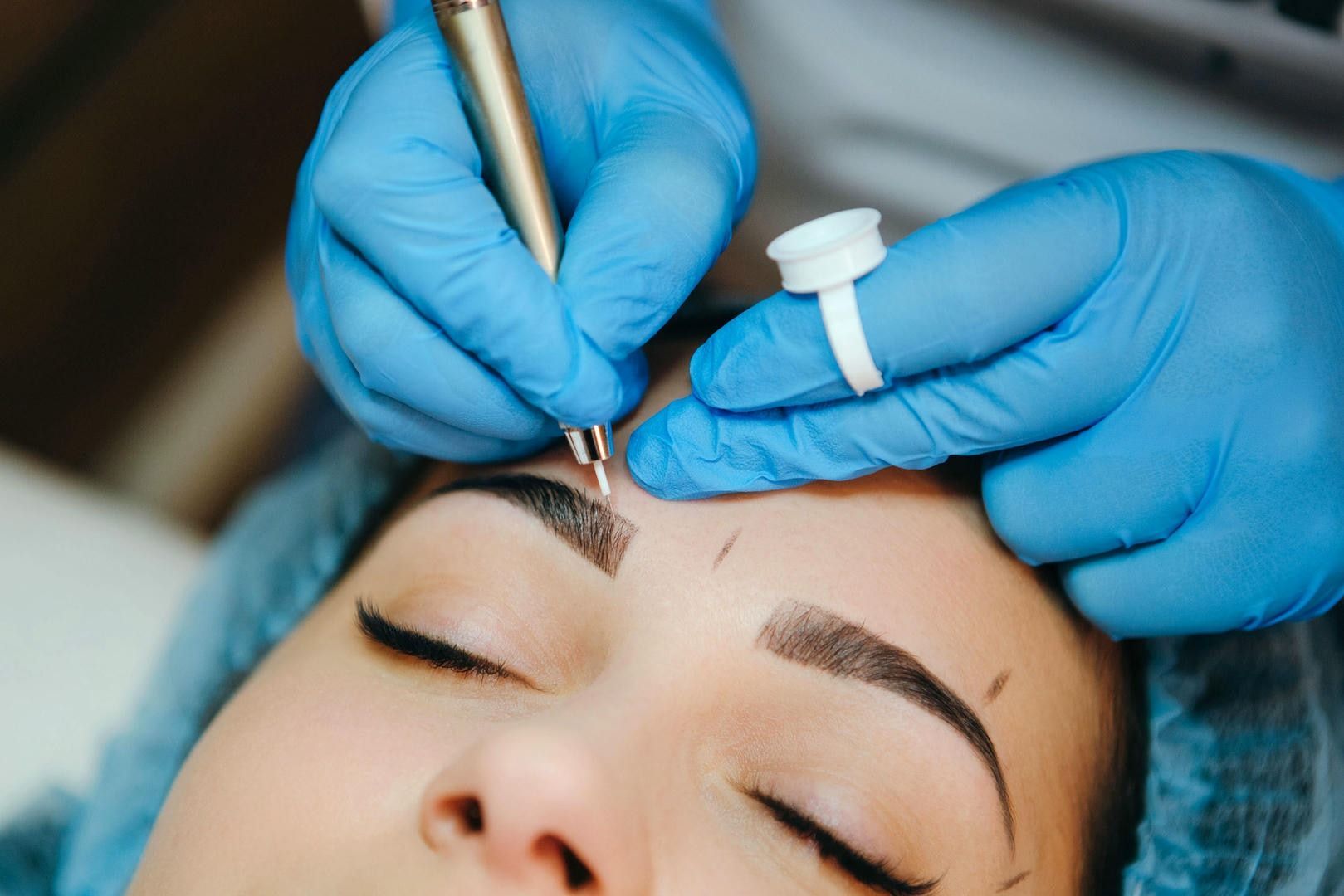 Person getting microblading; technician in blue gloves using pen to create eyebrow.