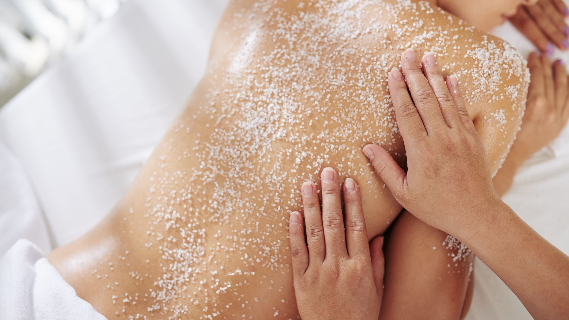 Woman receiving a back scrub at a spa, with white exfoliating product being massaged in by two hands.