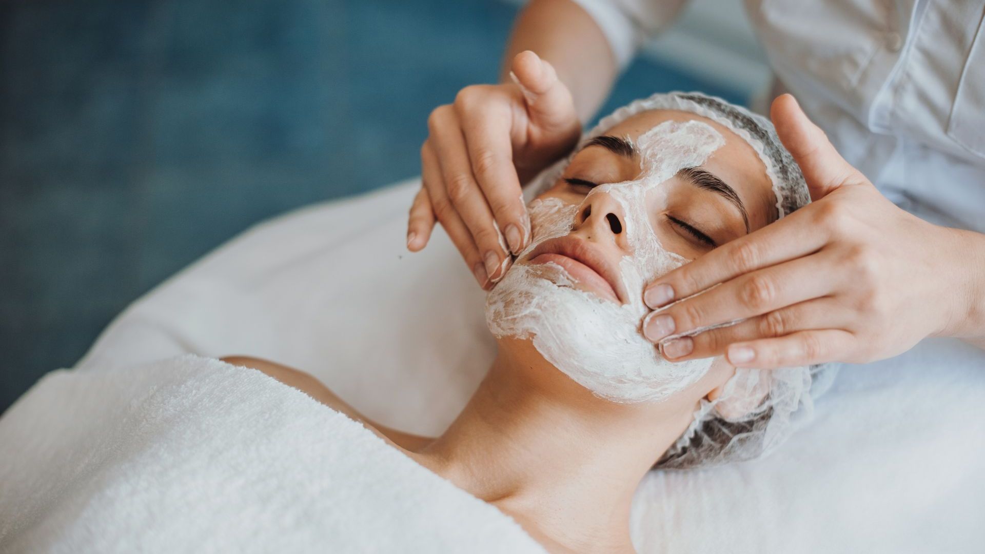 Woman receiving a facial at a spa, wearing a hair net and face mask; hands applying cream.