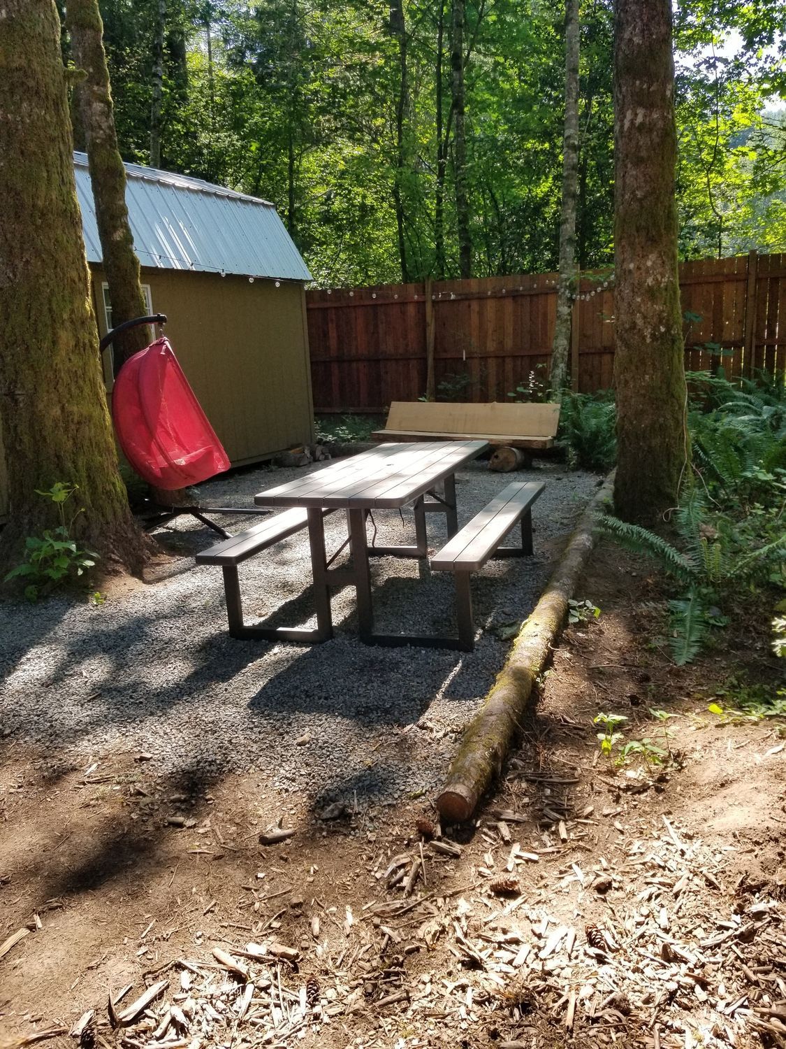 Wooded campsite with picnic table, red chair, small shed, and wooden fence in sunlight