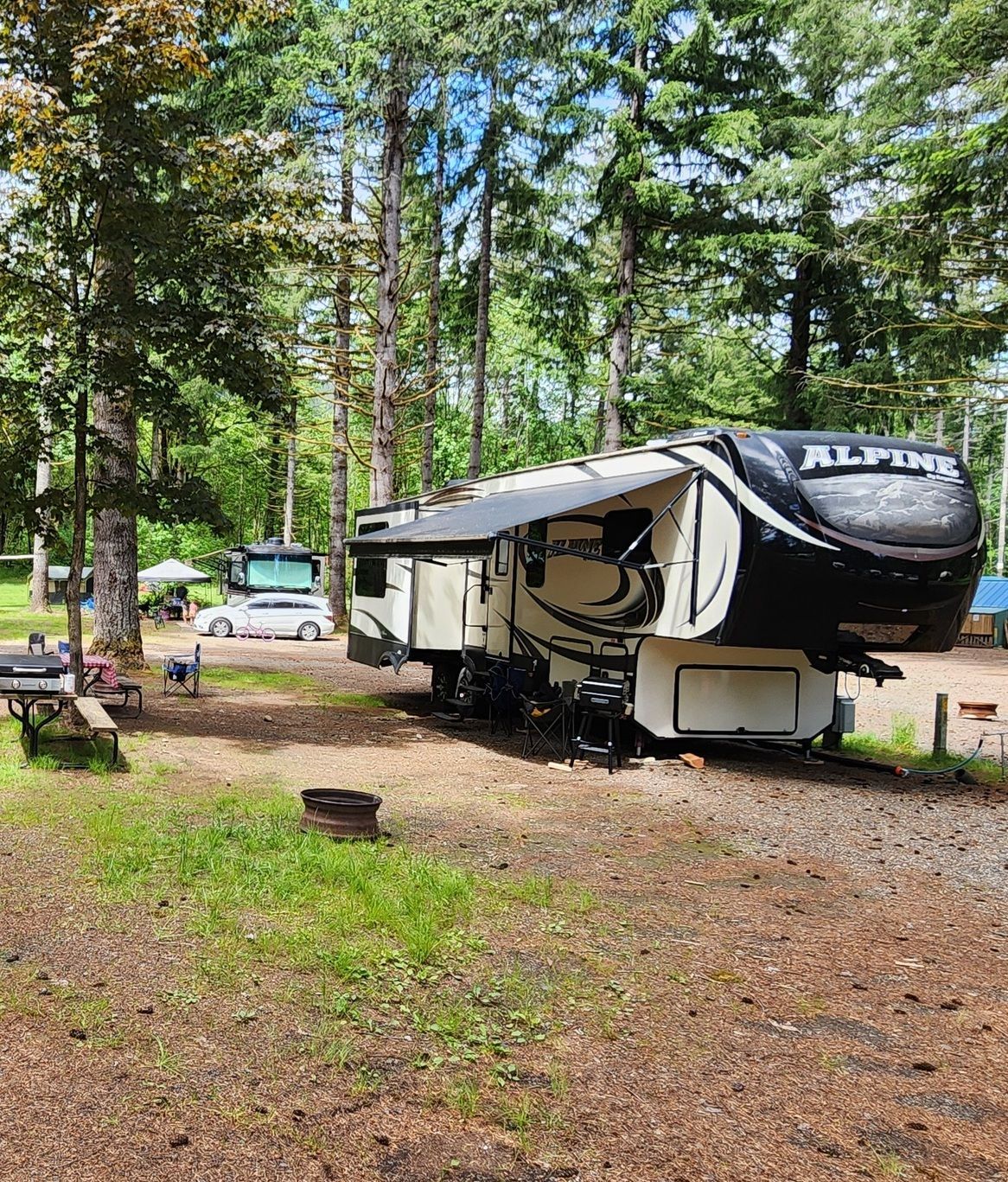 White and black ALP travel trailer parked in a wooded campsite beside a car and picnic area.