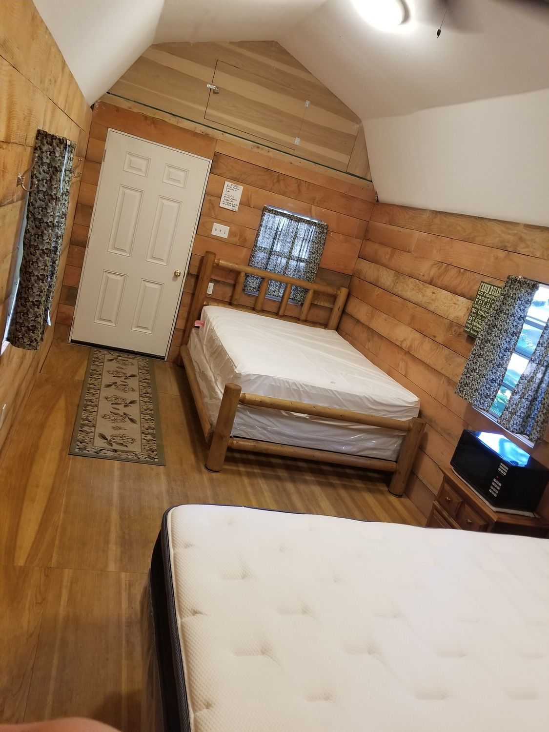 Rustic cabin bedroom with two beds, wood-paneled walls, white door, and small windows with curtains