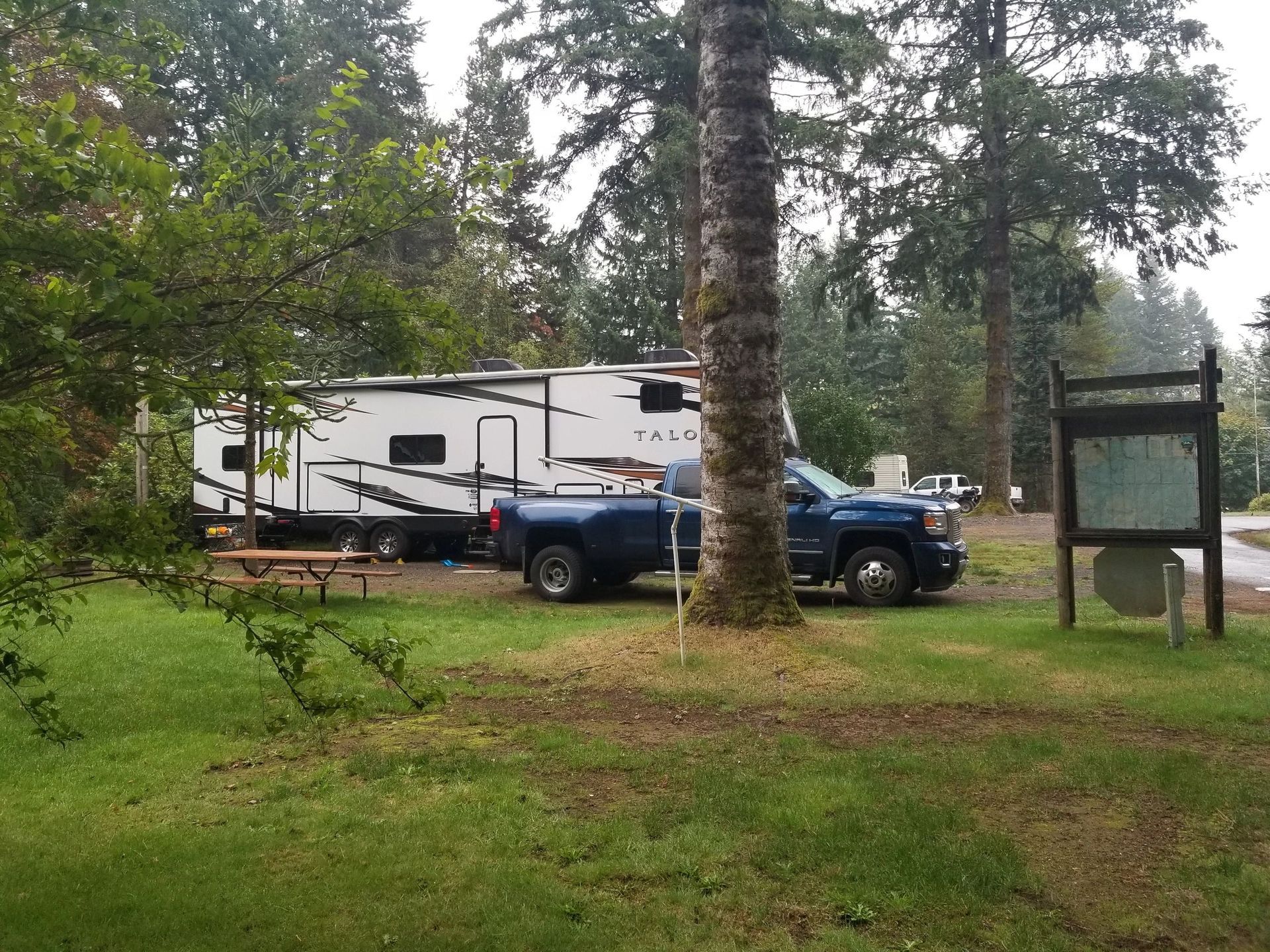 Blue pickup truck towing a white RV parked in a wooded campsite beside a signboard.