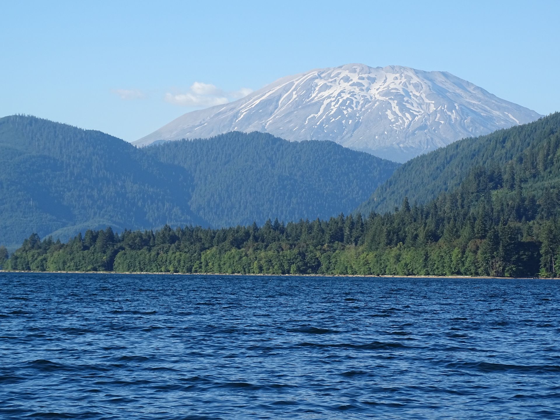 Snow-capped mountain above forested hills and a blue lake under a clear sky