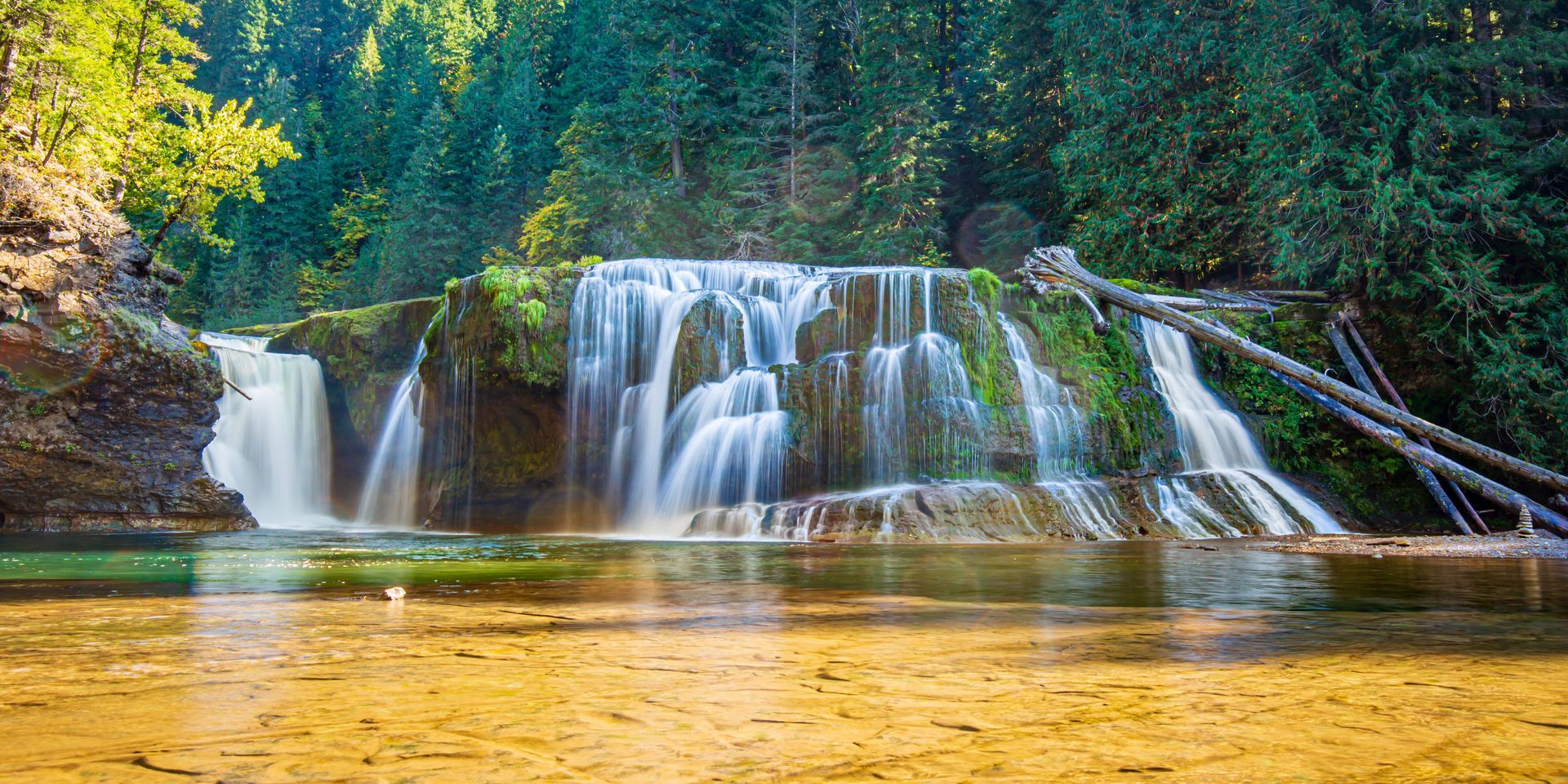 Wide waterfall cascading over rocky ledges into a calm amber pool, framed by lush green forest