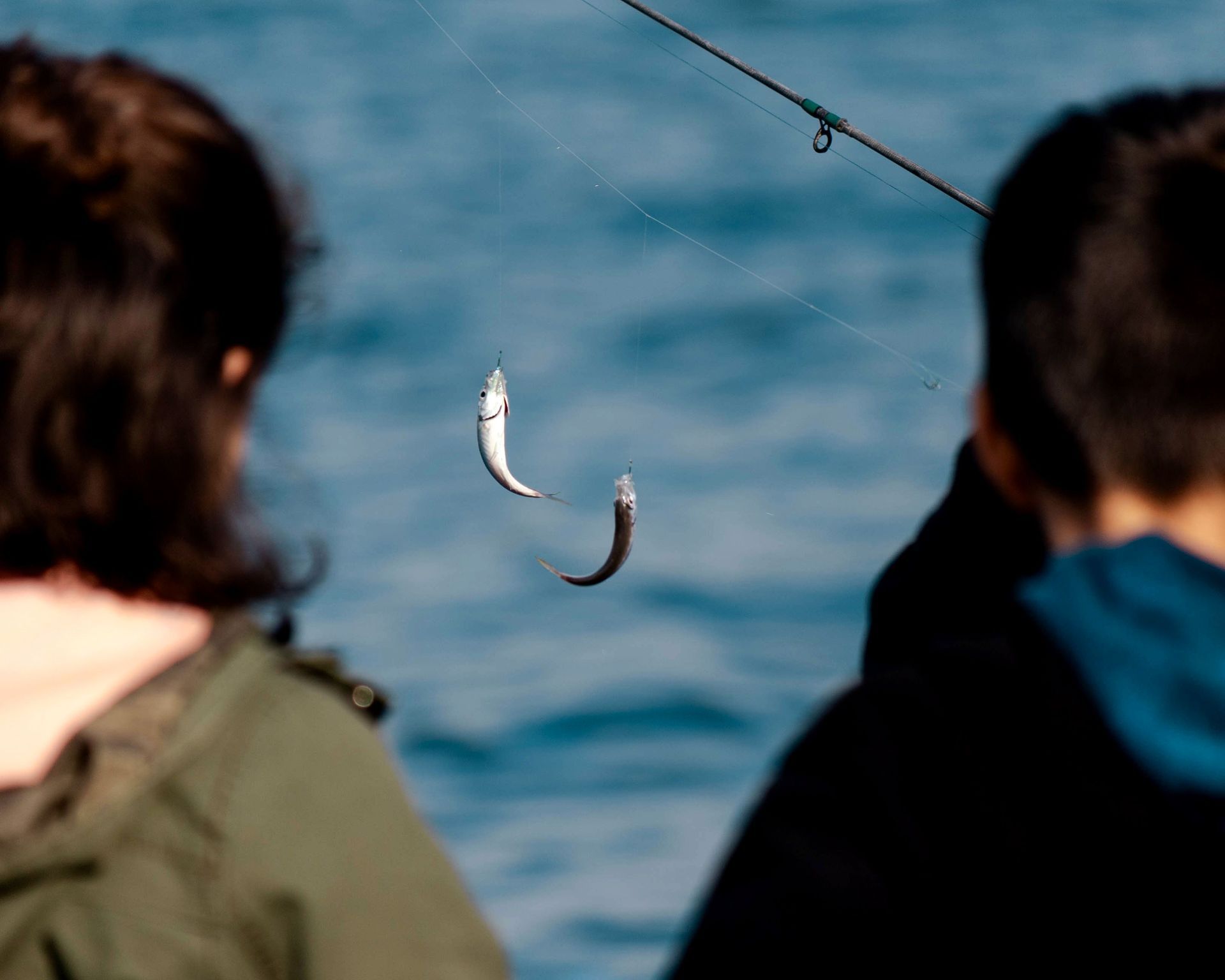 Two people watch a fishing line with two dangling fish over blue water.