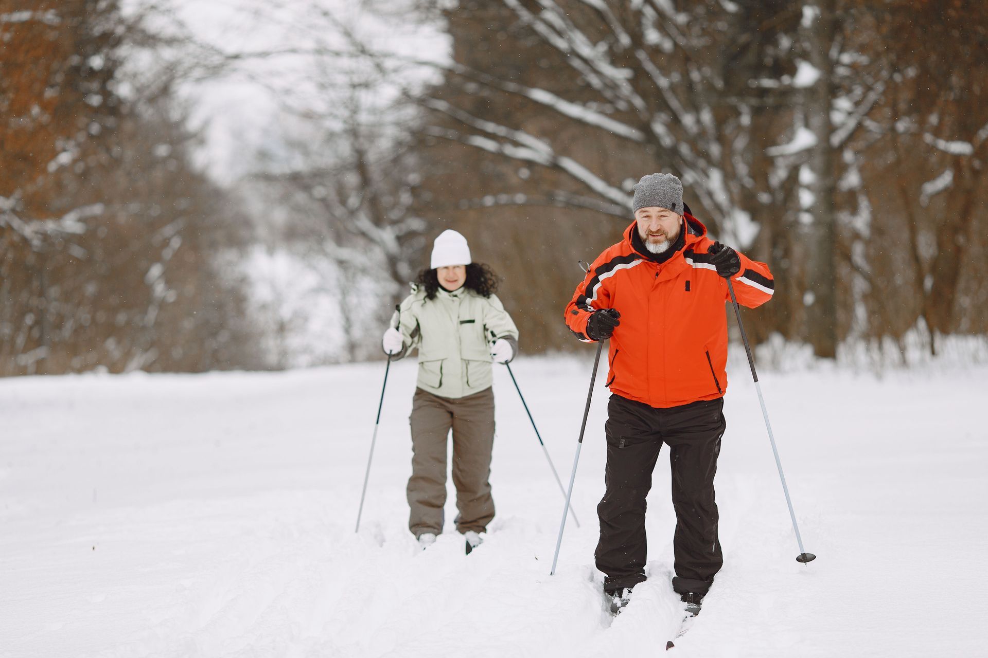 Two people cross-country skiing on a snowy trail, one in orange and one in white.