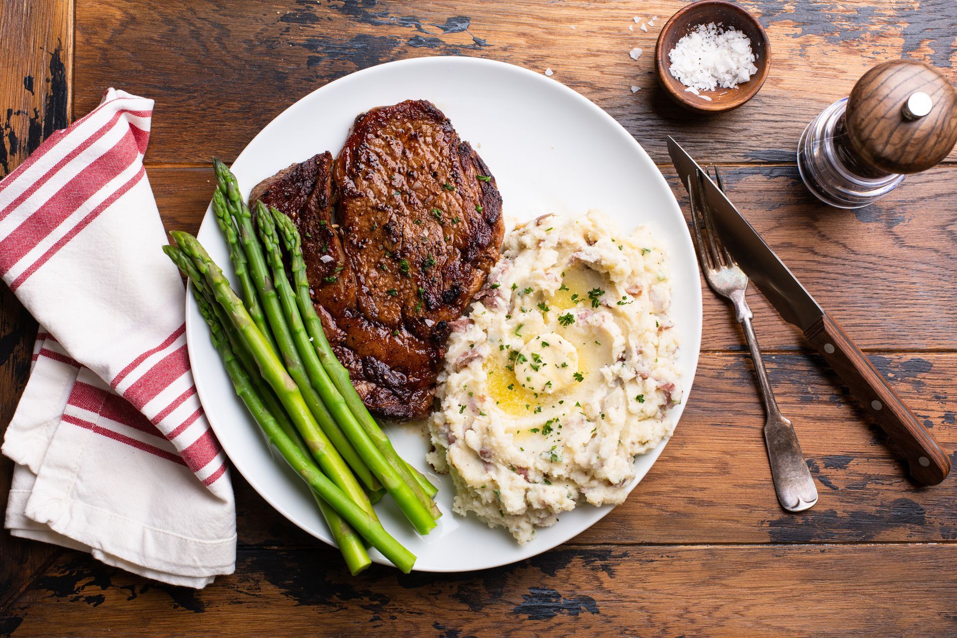 Steak, asparagus, and mashed potatoes on a white plate on a rustic wooden table with napkin and cutlery