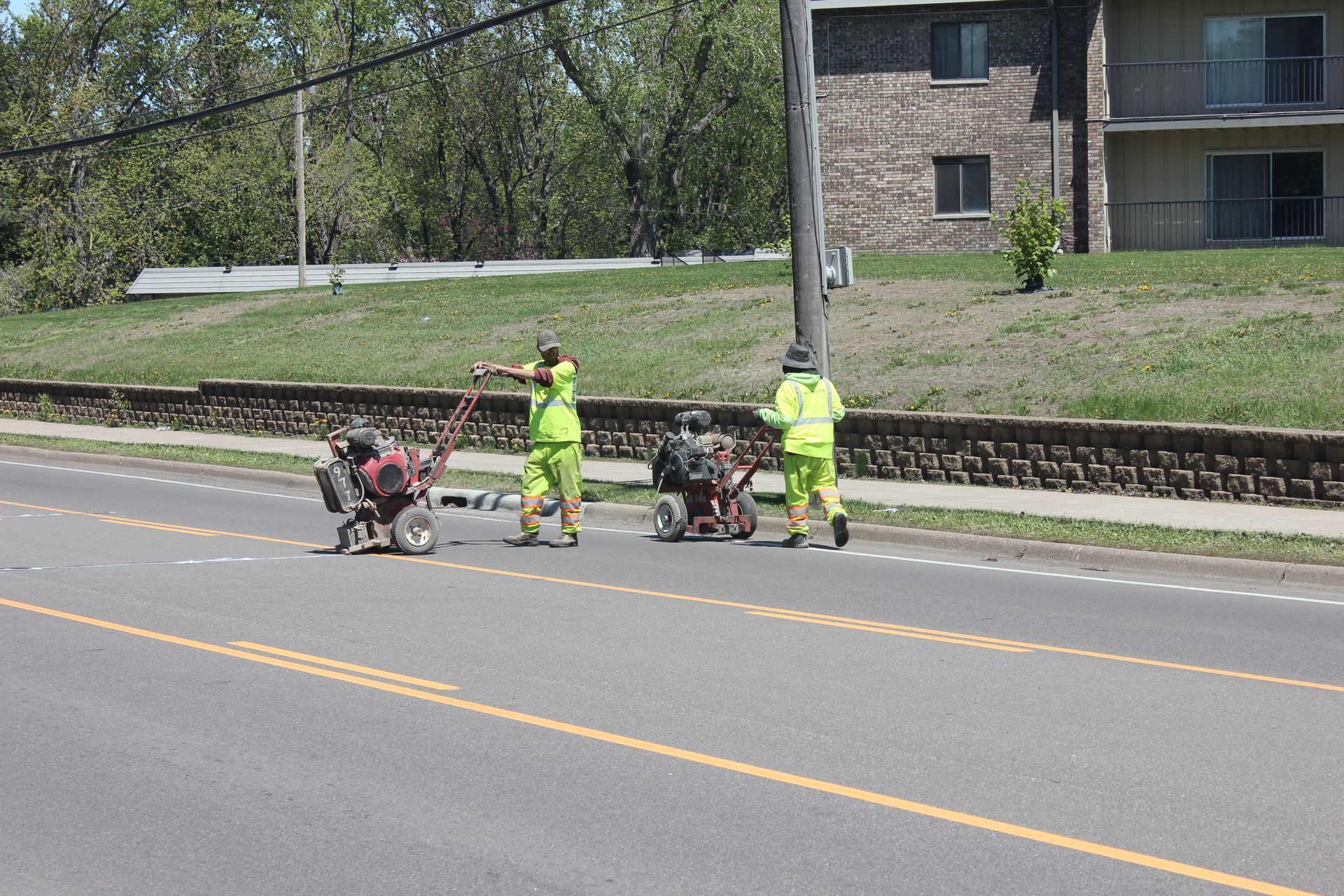 Two road workers in neon vests paint yellow lines on a street, near a stone wall and building.