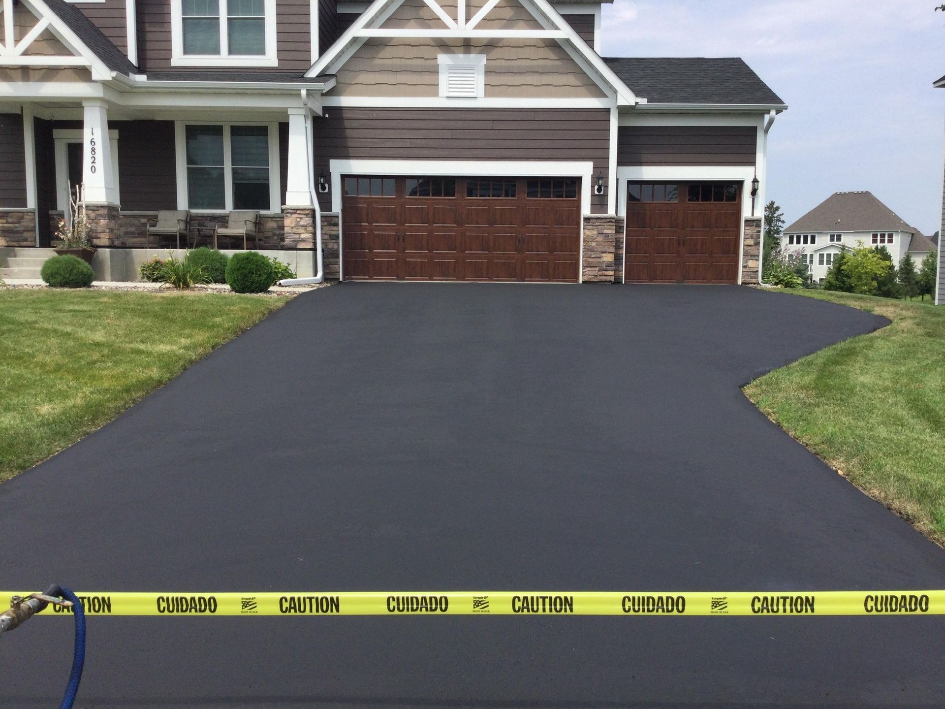 Newly paved black asphalt driveway to a brown-trimmed house with a two-car garage, yellow caution tape stretched across.