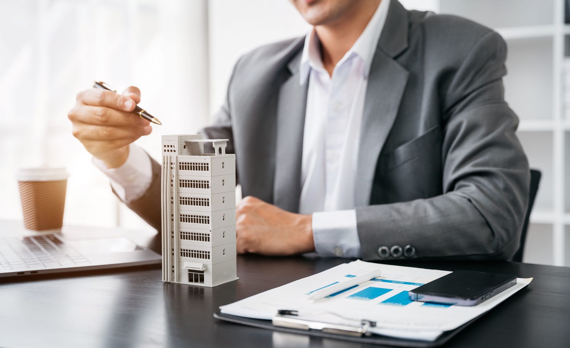 Man in suit pointing at a model building, documents and phone on desk.