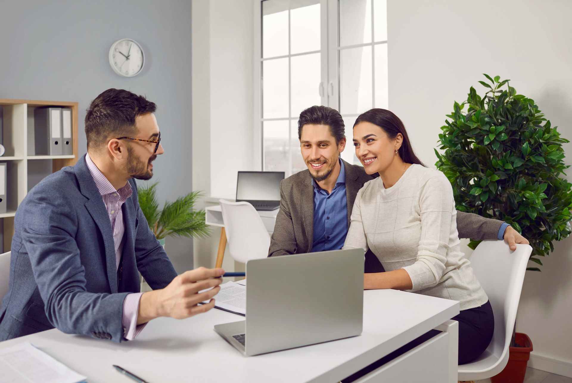 Real estate advisor in glasses showing laptop to smiling couple at a desk in an office.