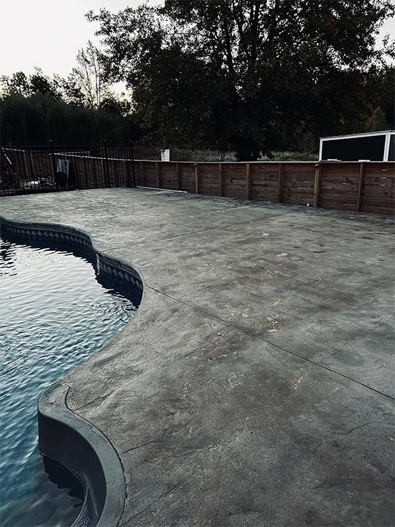 A concrete patio next to a swimming pool with trees in the background.