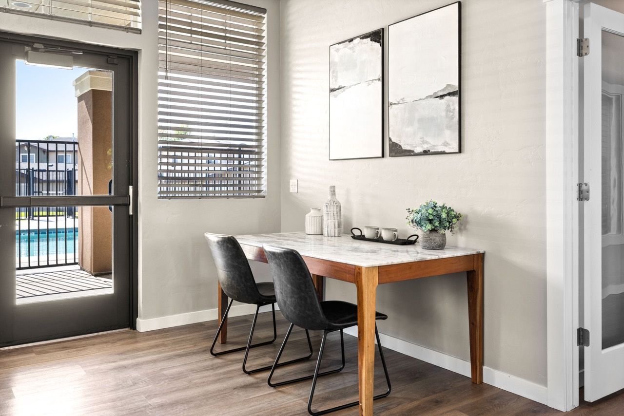 Dining area in an apartment with a marble table, two chairs, blinds, and a balcony door.
