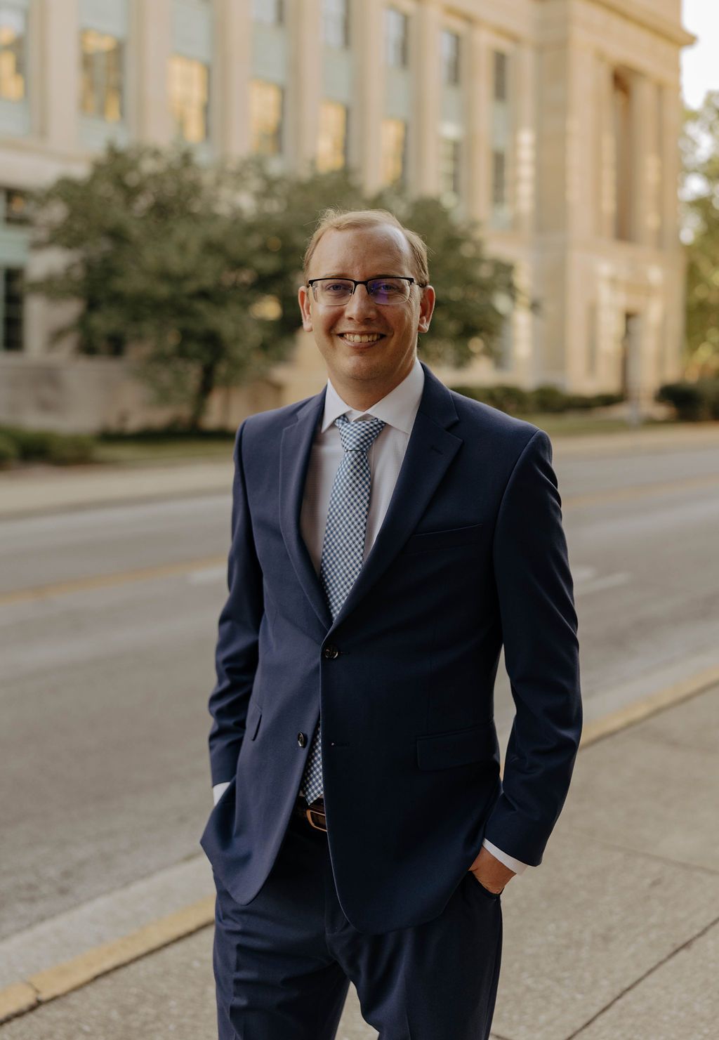 Thom in a blue suit and tie is standing on the sidewalk with his hands in his pockets.