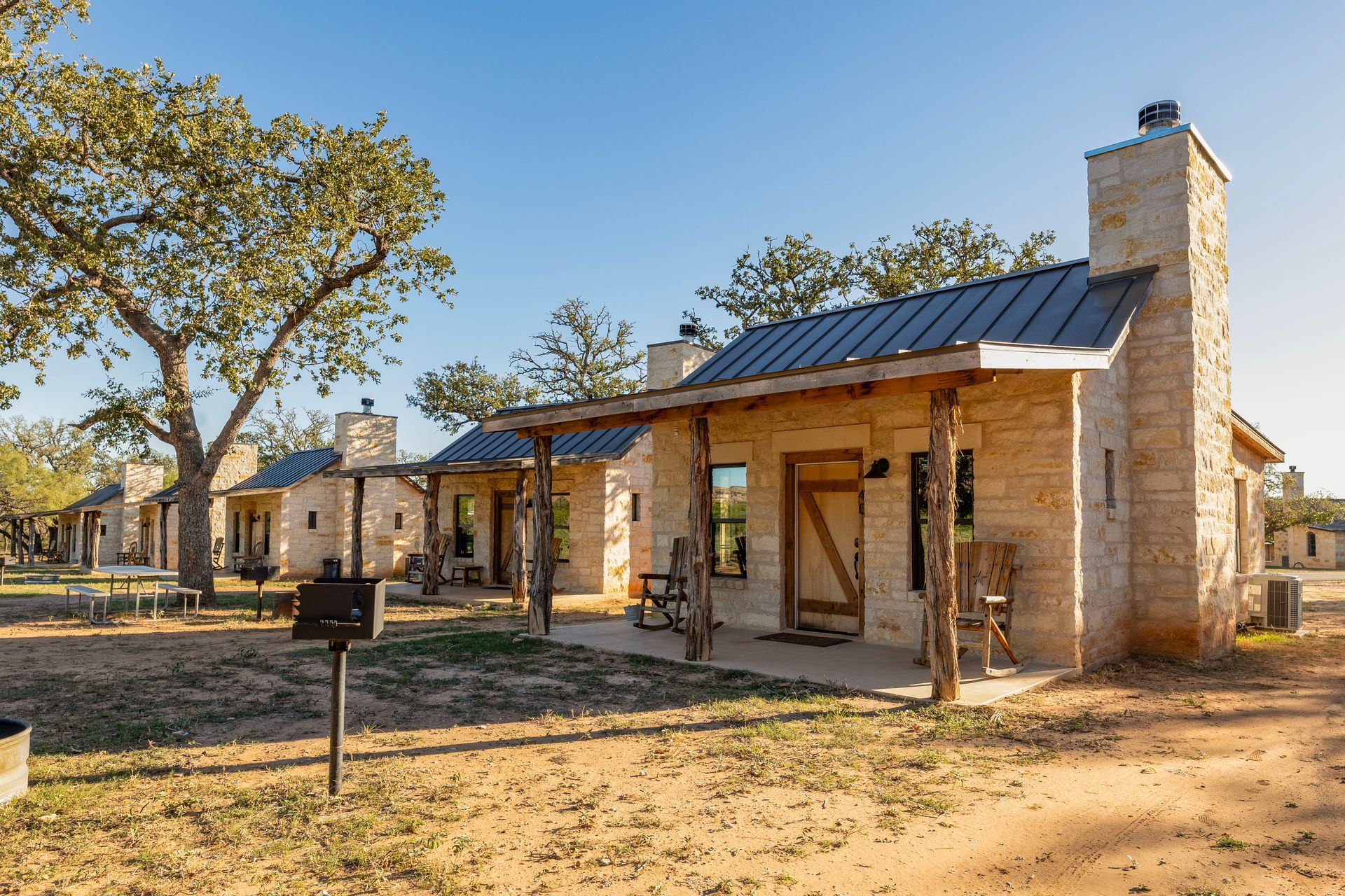 Stone cabins with porches under a blue sky. Rural setting, wooden posts, and a metal roof.