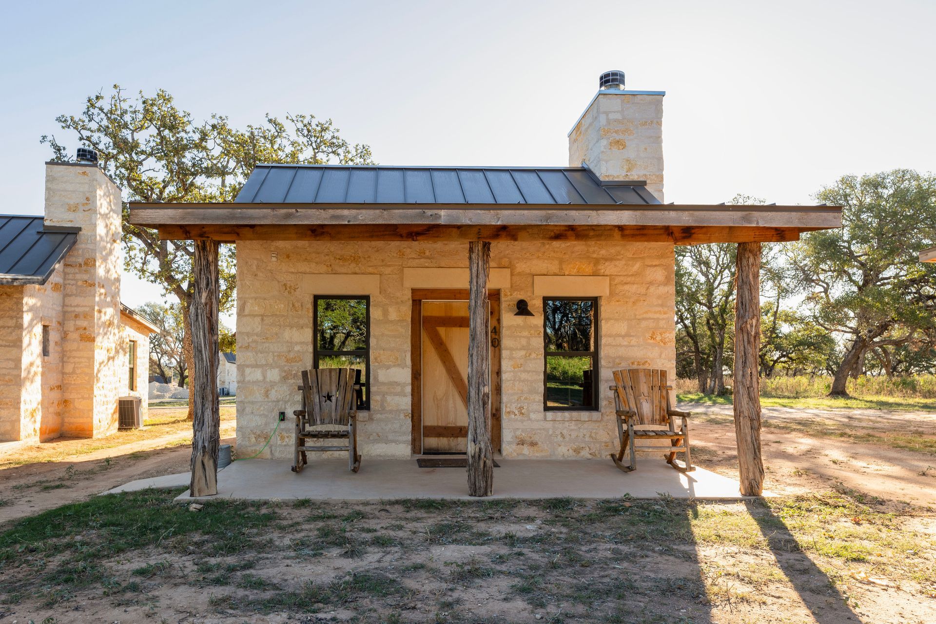 Stone cottage with porch, rocking chairs, wooden door, and metal roof. Set in a grassy field.