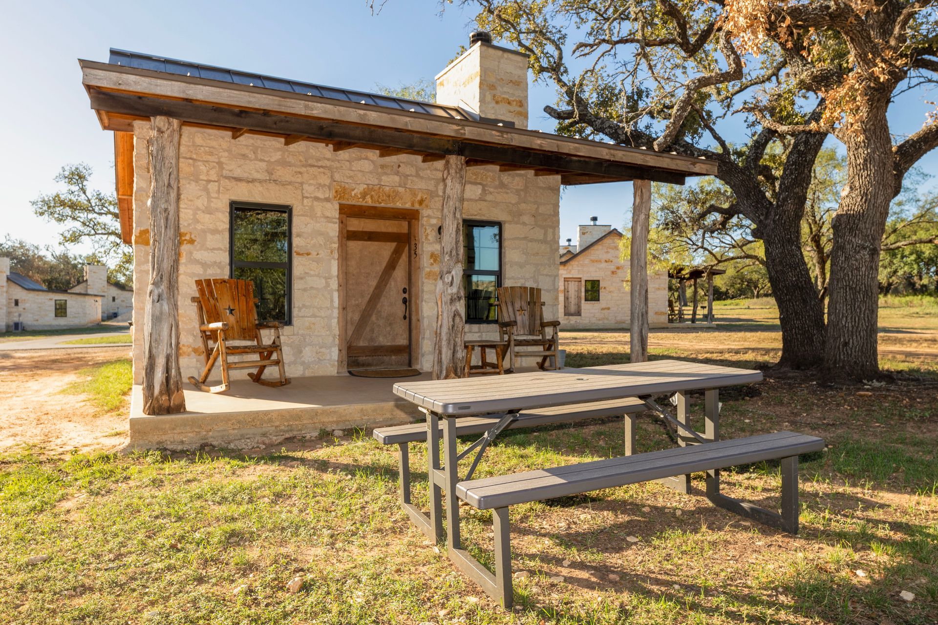 Small stone cabin with porch, chairs, and picnic table on grassy land, under a tree.