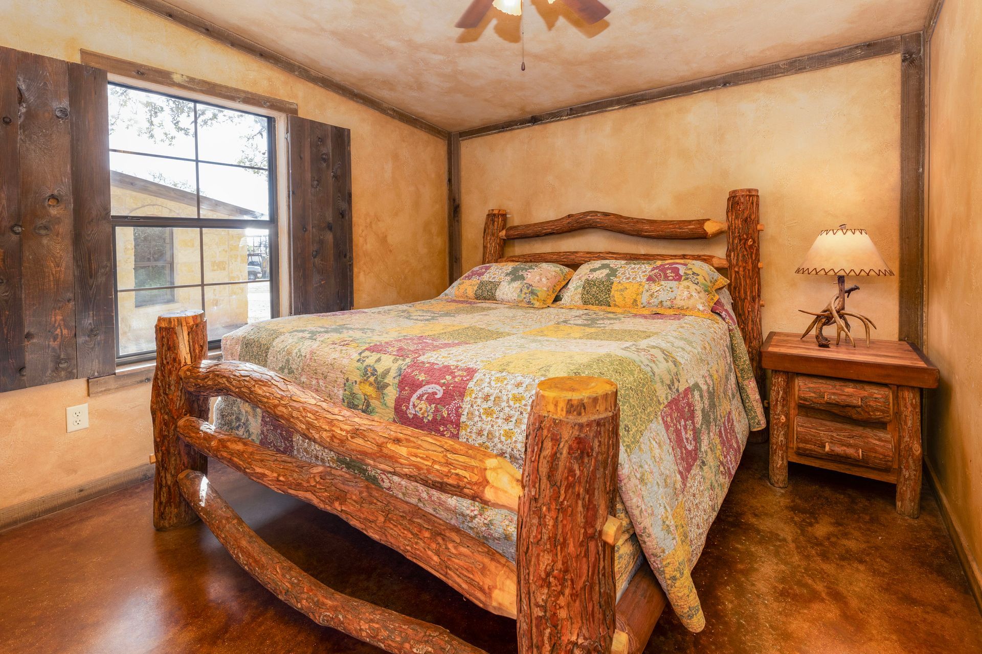 Rustic bedroom with wooden bed, quilt, nightstand, and window with shutters.