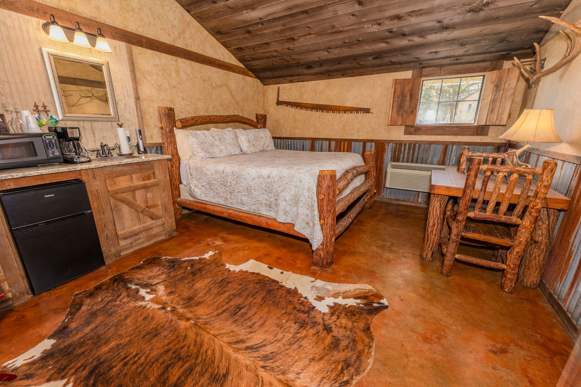 Rustic bedroom with a bed, desk, mini-fridge, and cowhide rug. Wooden beams and antlers add to the decor.