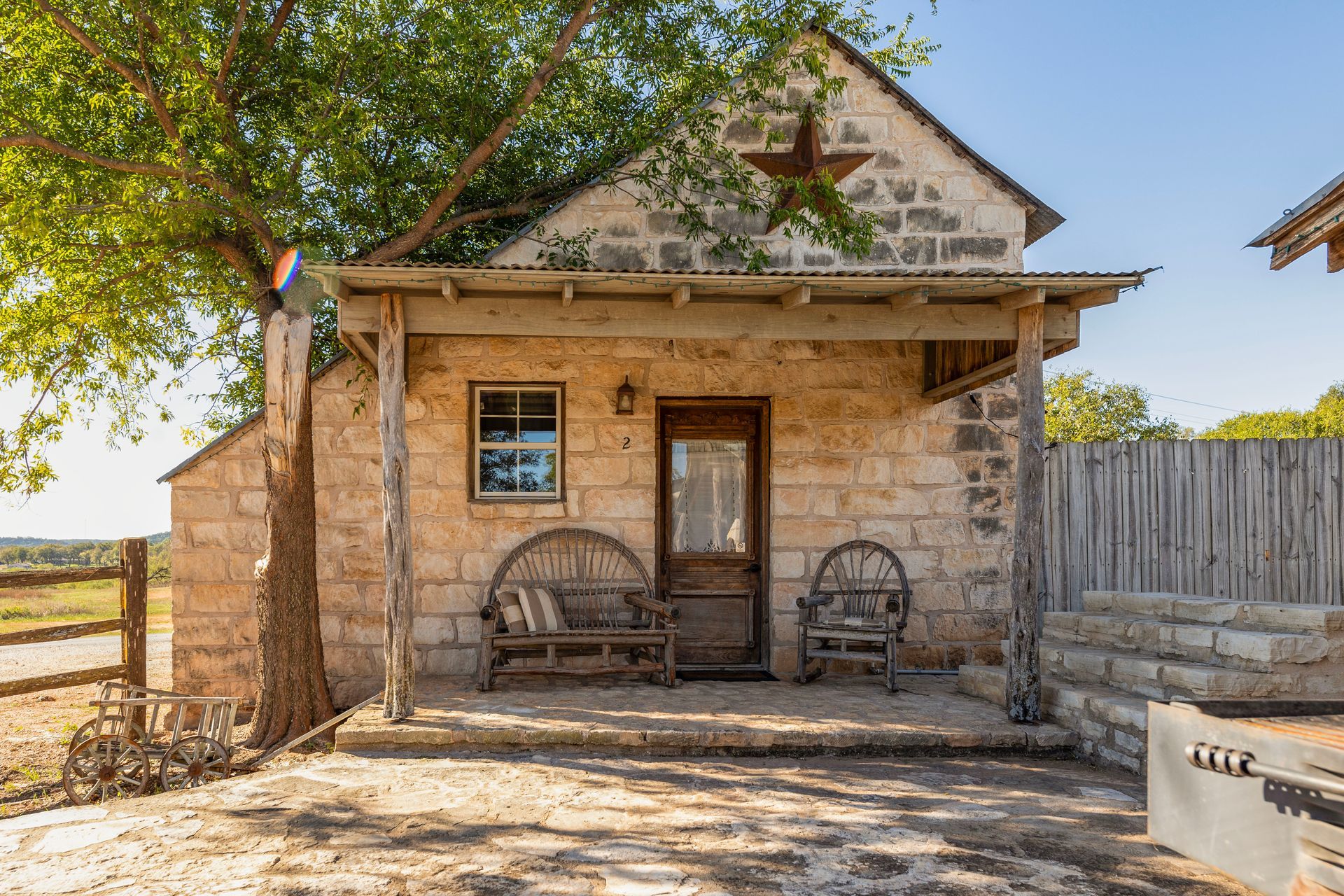 Stone cottage with covered porch, star decor, benches, and a grill, in a sunny outdoor setting.