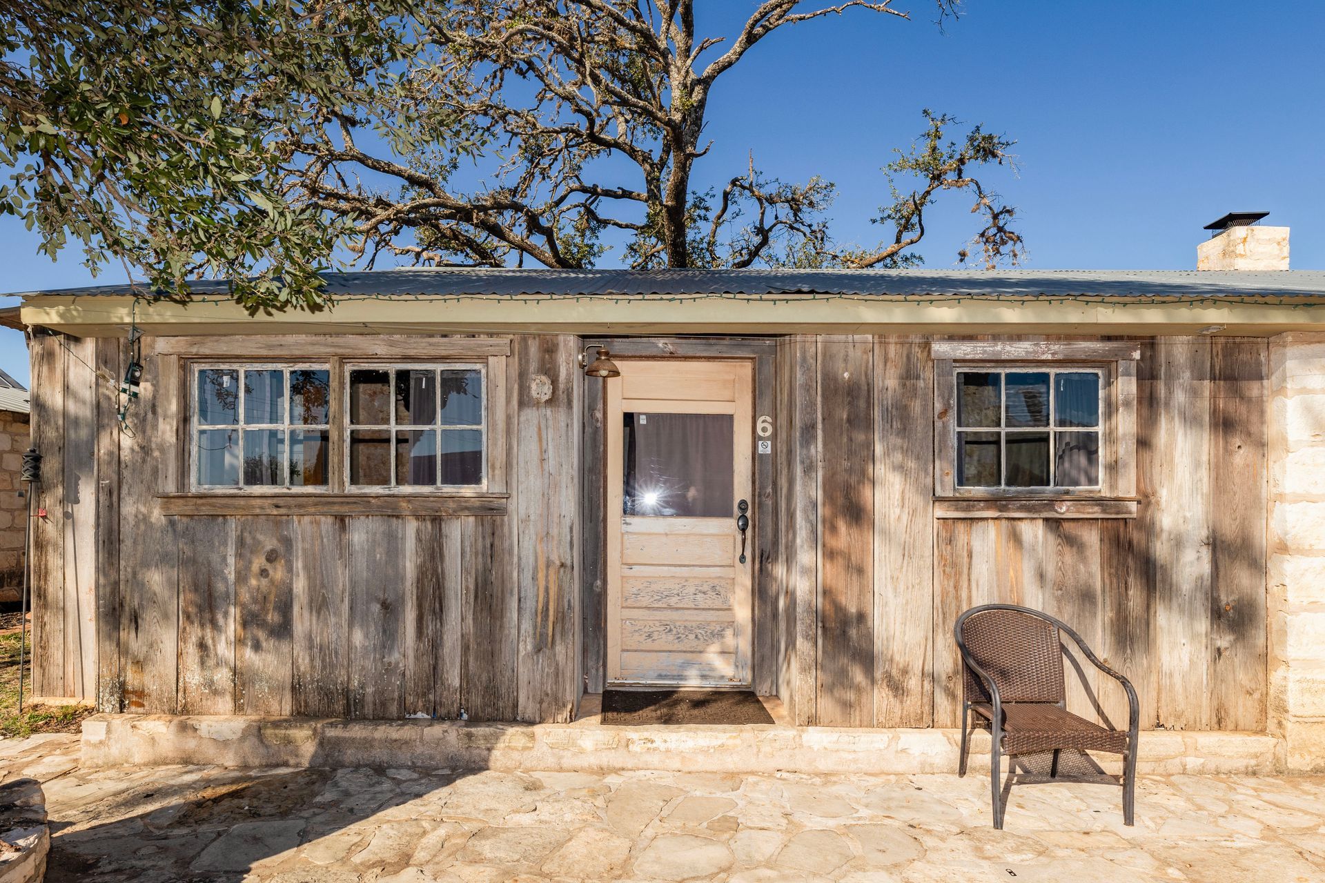 Rustic wooden cabin with two windows, a door, and a chair on a stone patio under a sunny sky.