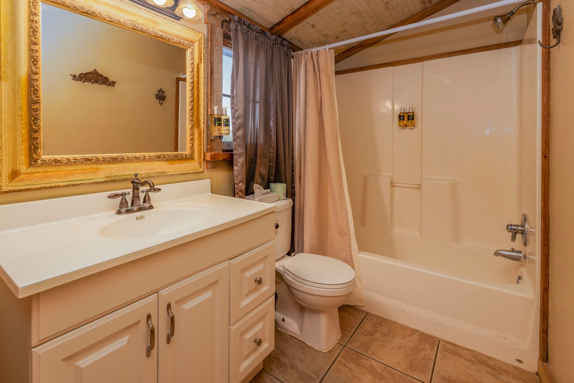 Bathroom with white vanity, toilet, and shower. Beige walls and flooring. Gold-framed mirror and tan shower curtain.