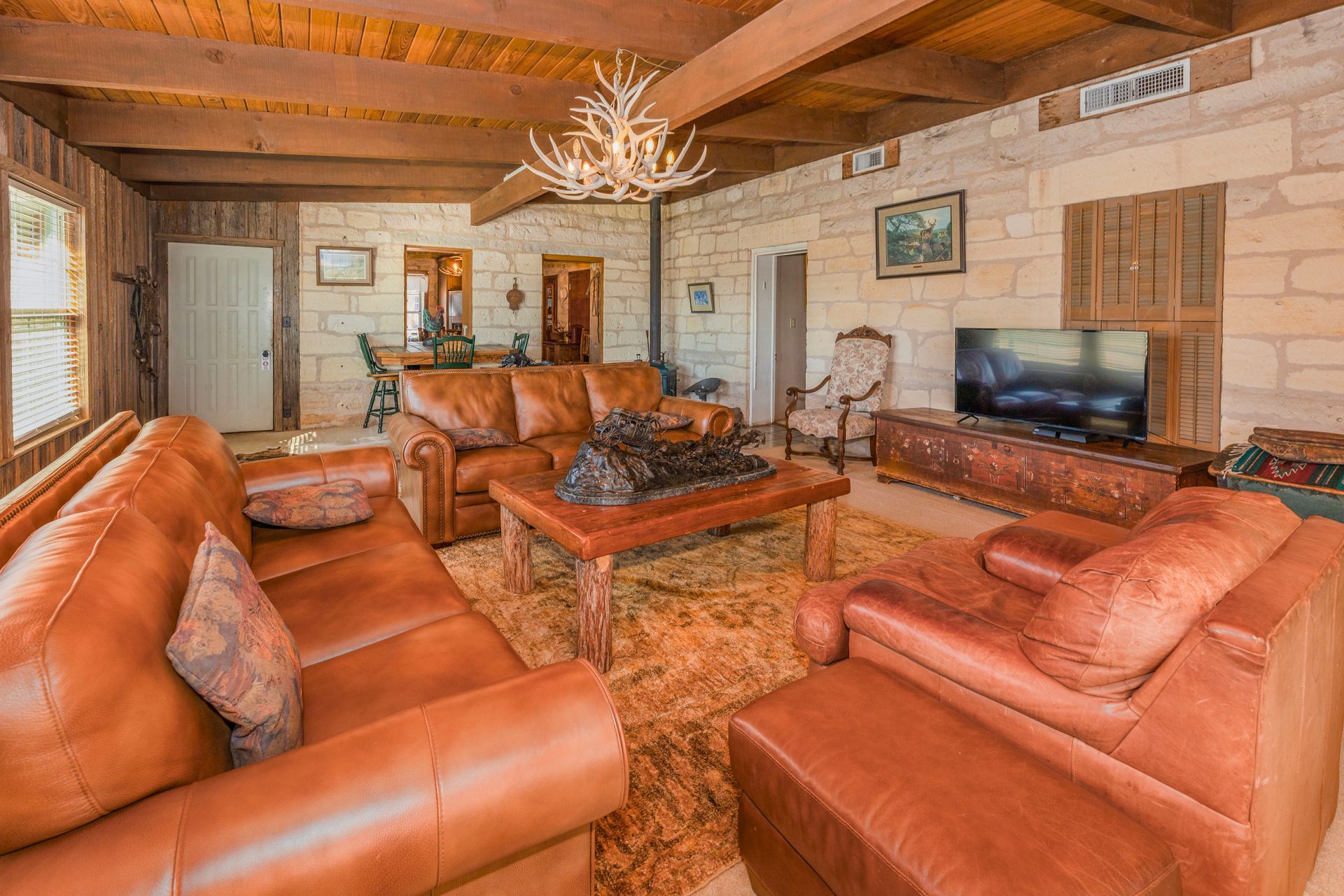 Living room with leather furniture, stone walls, wood beams, and a chandelier.