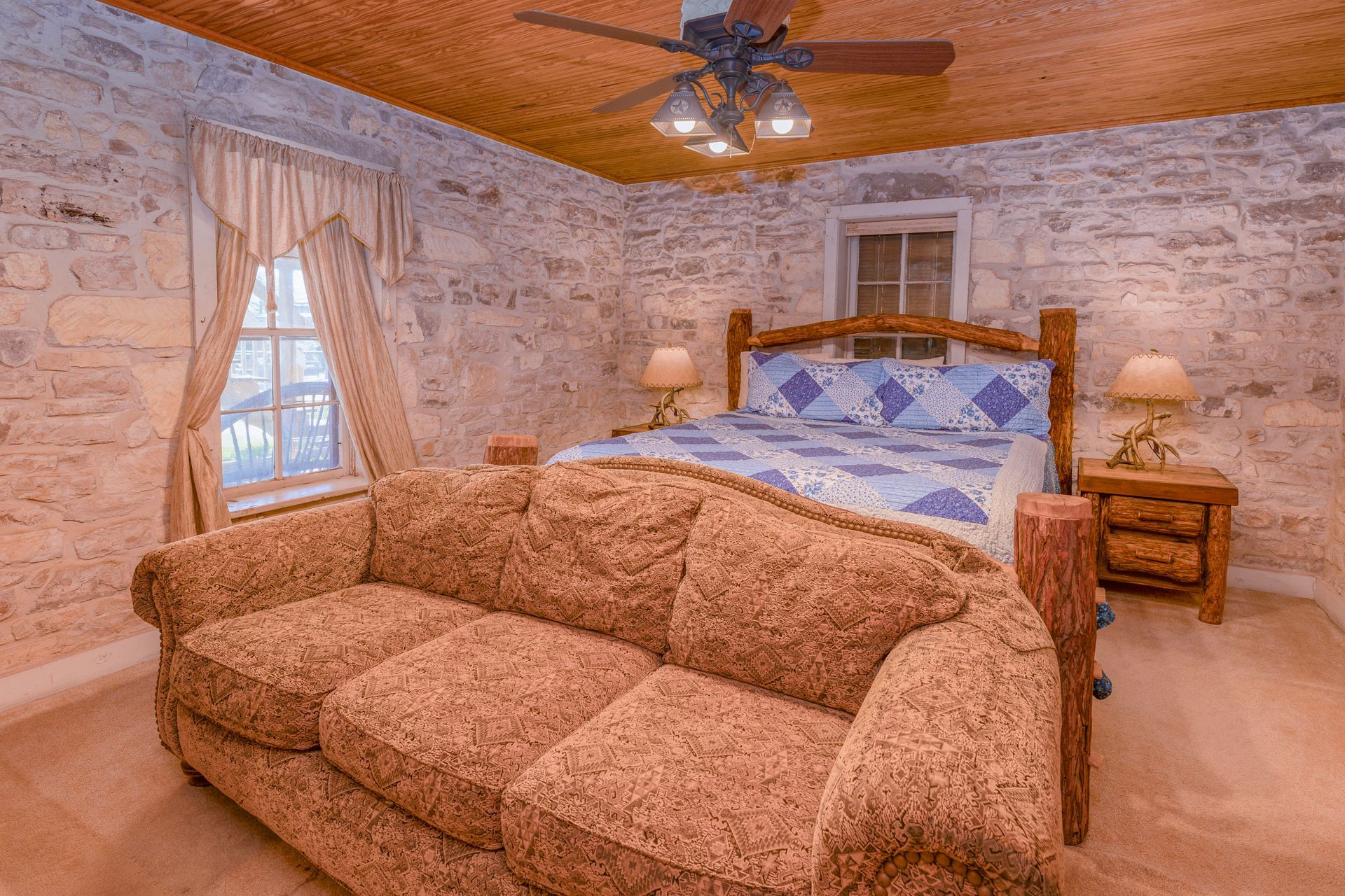 Cozy bedroom with a stone wall and wooden ceiling; a bed, sofa, and window are visible.
