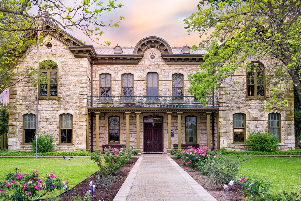 Historic two-story stone building with symmetrical design, walkway, and flowers, under a pastel sky.
