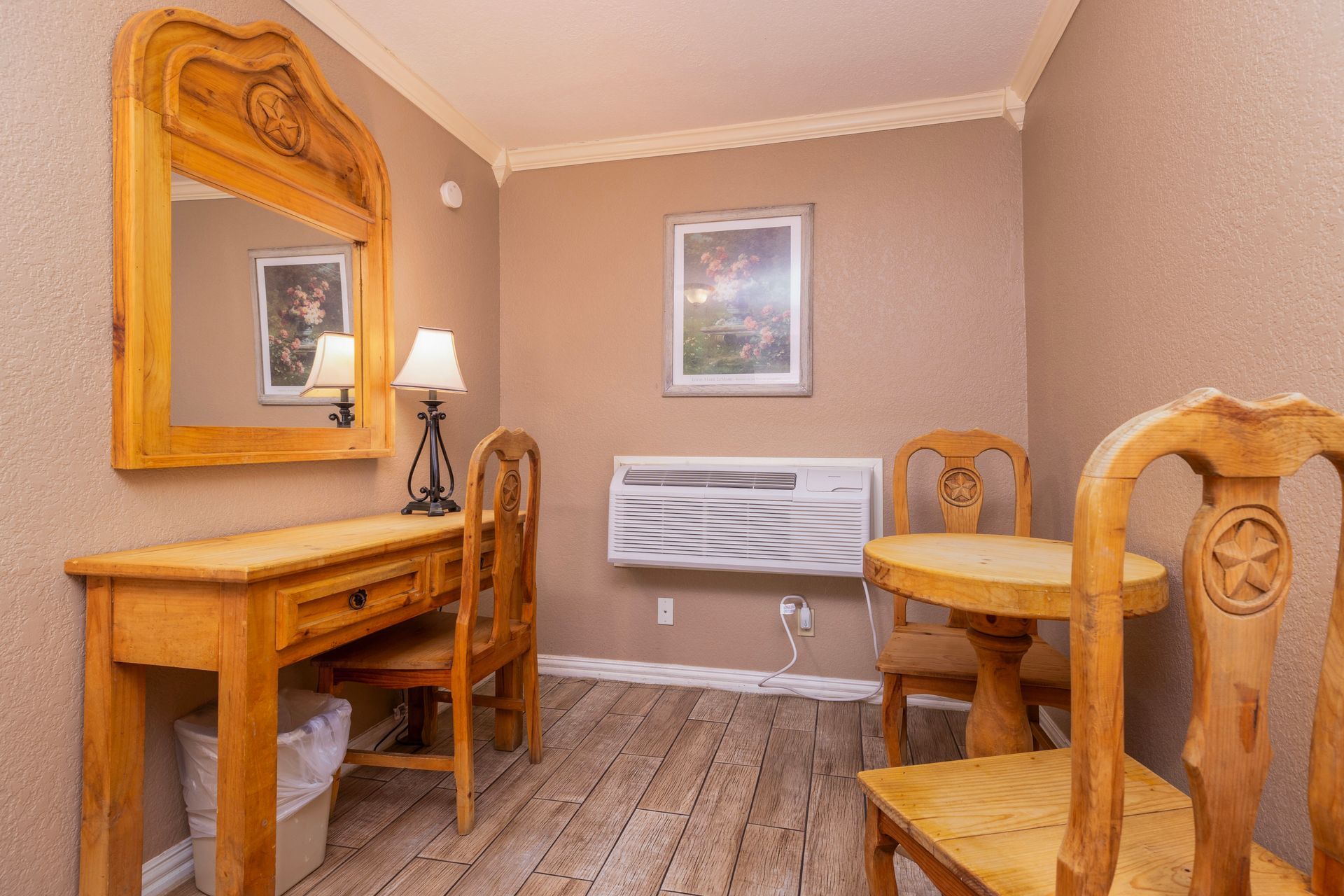 Hotel room with a desk, mirror, two chairs, and a small table. Soft lighting, beige walls, and wood floors.