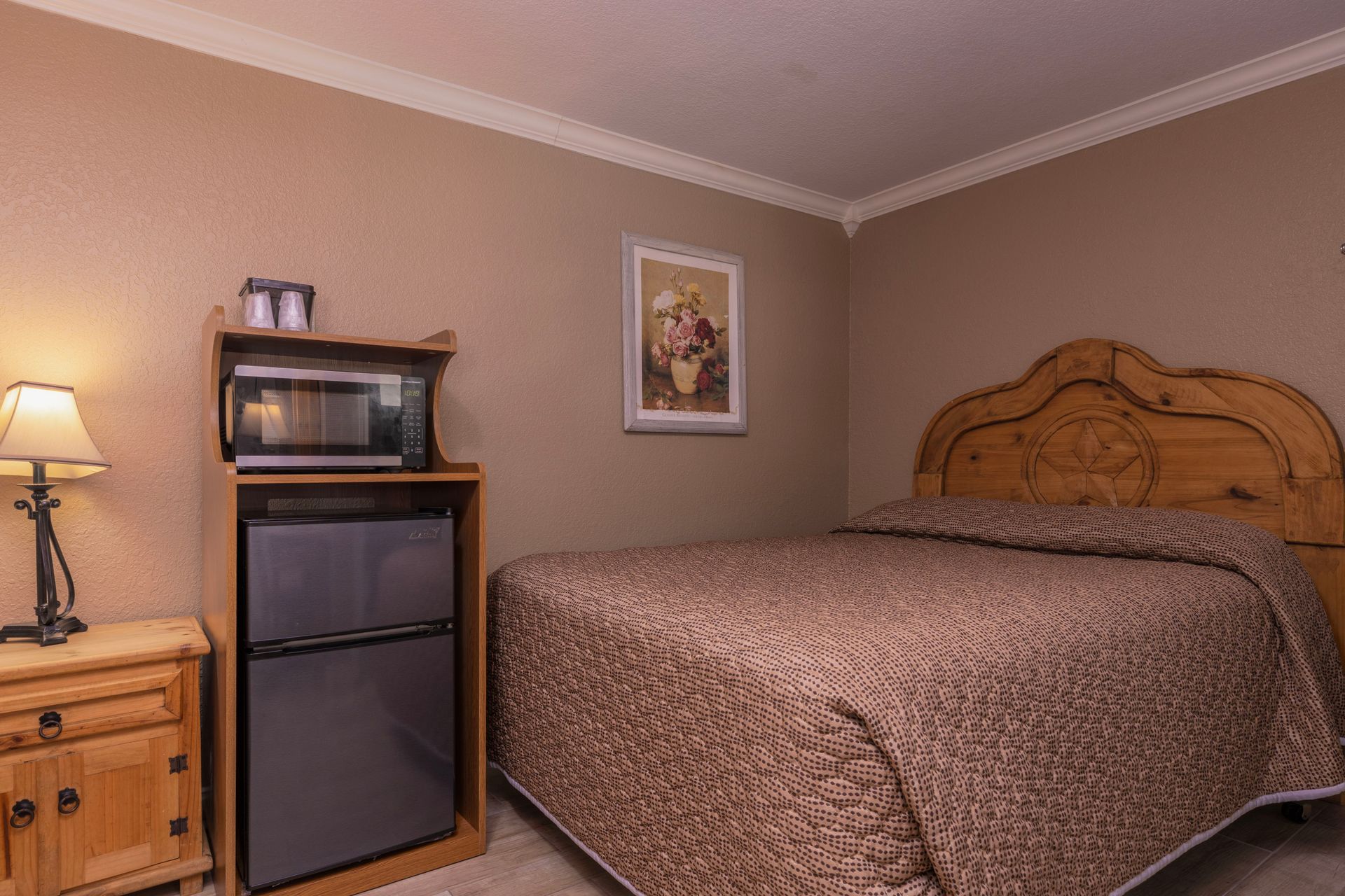 A hotel room with a bed, microwave, mini-fridge, and nightstand with a lamp. Brown and beige color scheme.