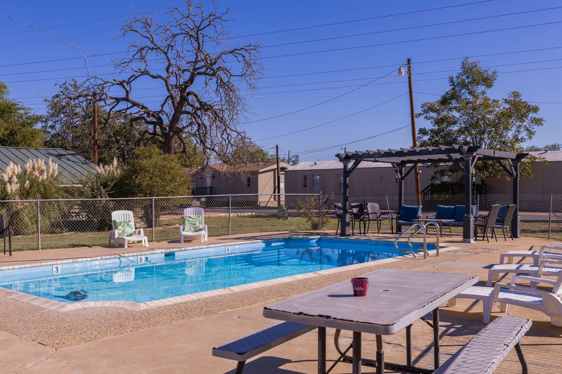 Pool area with picnic tables, chairs, and a pergola under a blue sky.