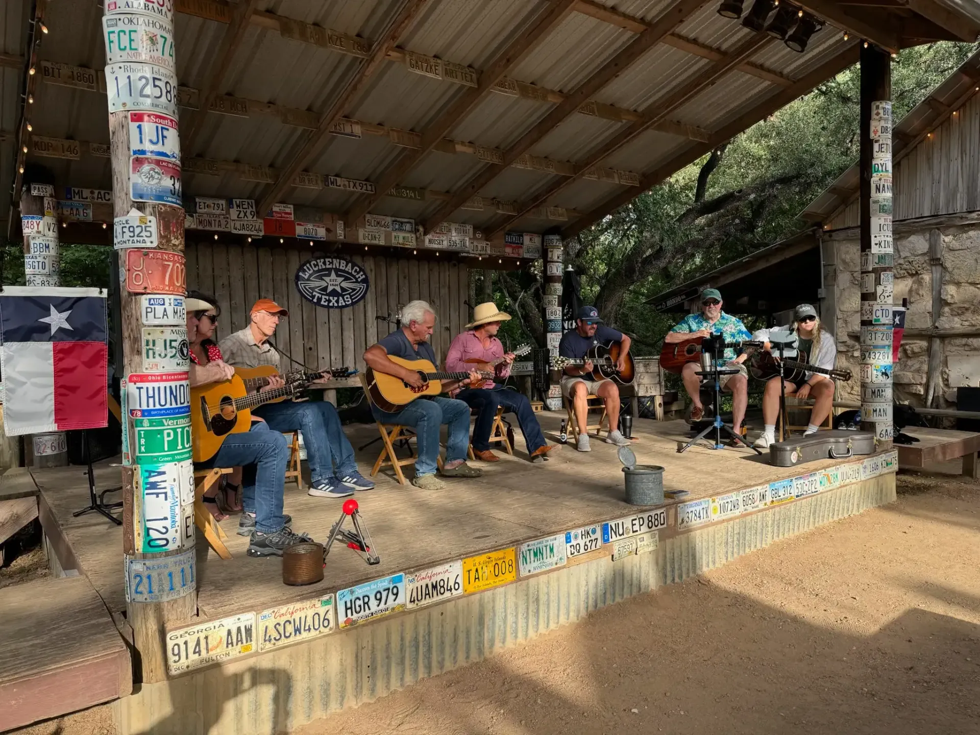 Six musicians with acoustic guitars perform on an outdoor stage decorated with license plates under a rustic shed roof.