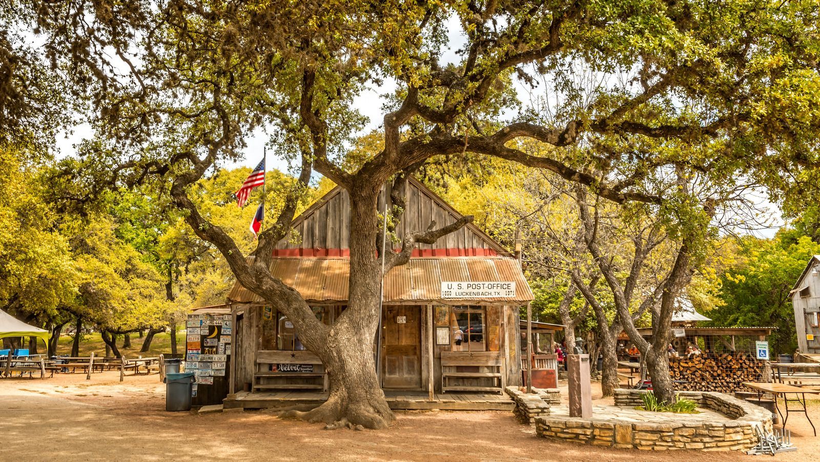 A weathered wooden building shaded by large oak trees, with an American flag displayed and rustic outdoor seating.