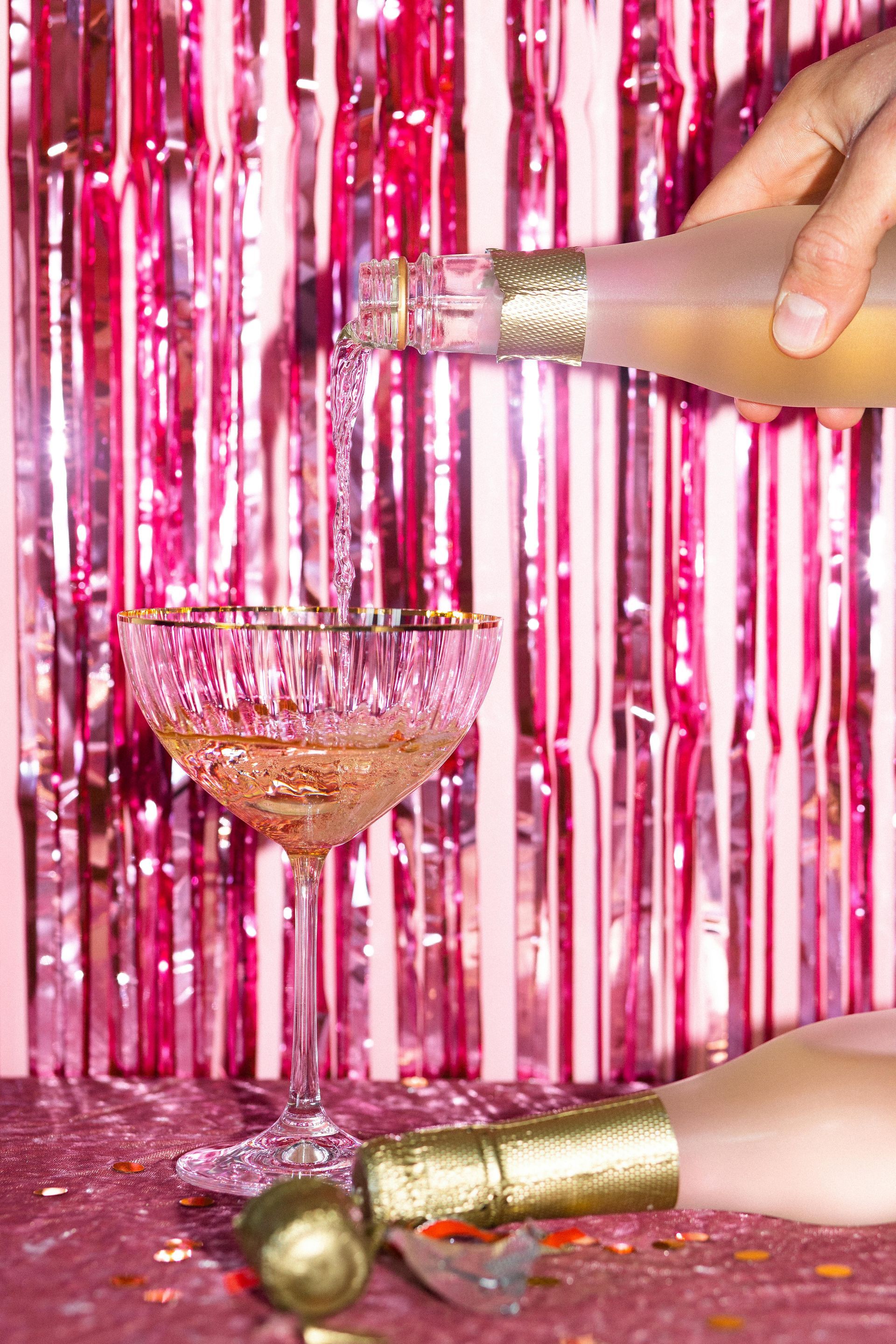 Champagne being poured into a glass. Pink background, bottle, glass, and confetti.
