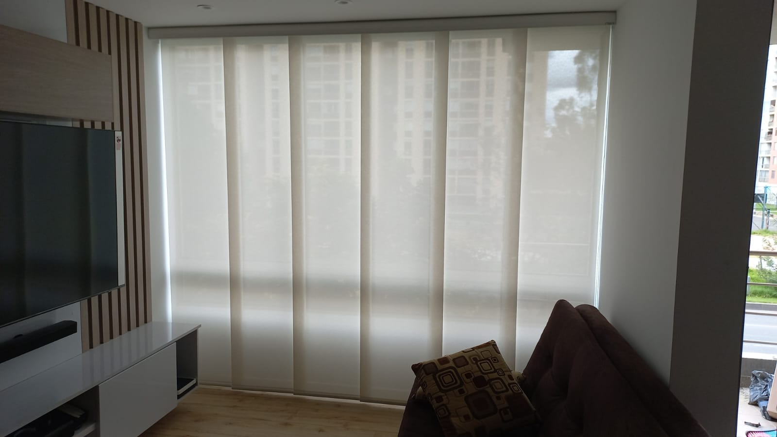 Salón con persianas verticales, televisor grande en la pared. Sofá y ventana.