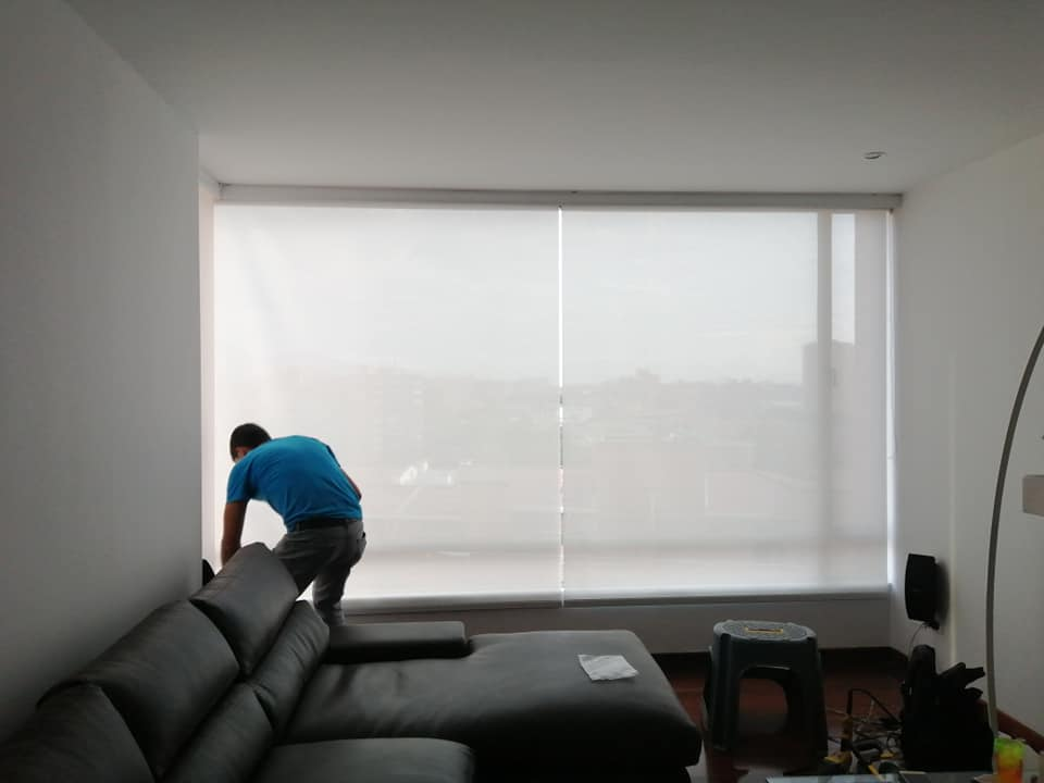 Persona ajustando las persianas en una sala de estar con paredes blancas, sofá gris y un taburete.