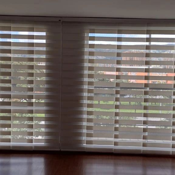 Persianas blancas delante de las ventanas, con la luz del sol creando sombras sobre el suelo de madera.