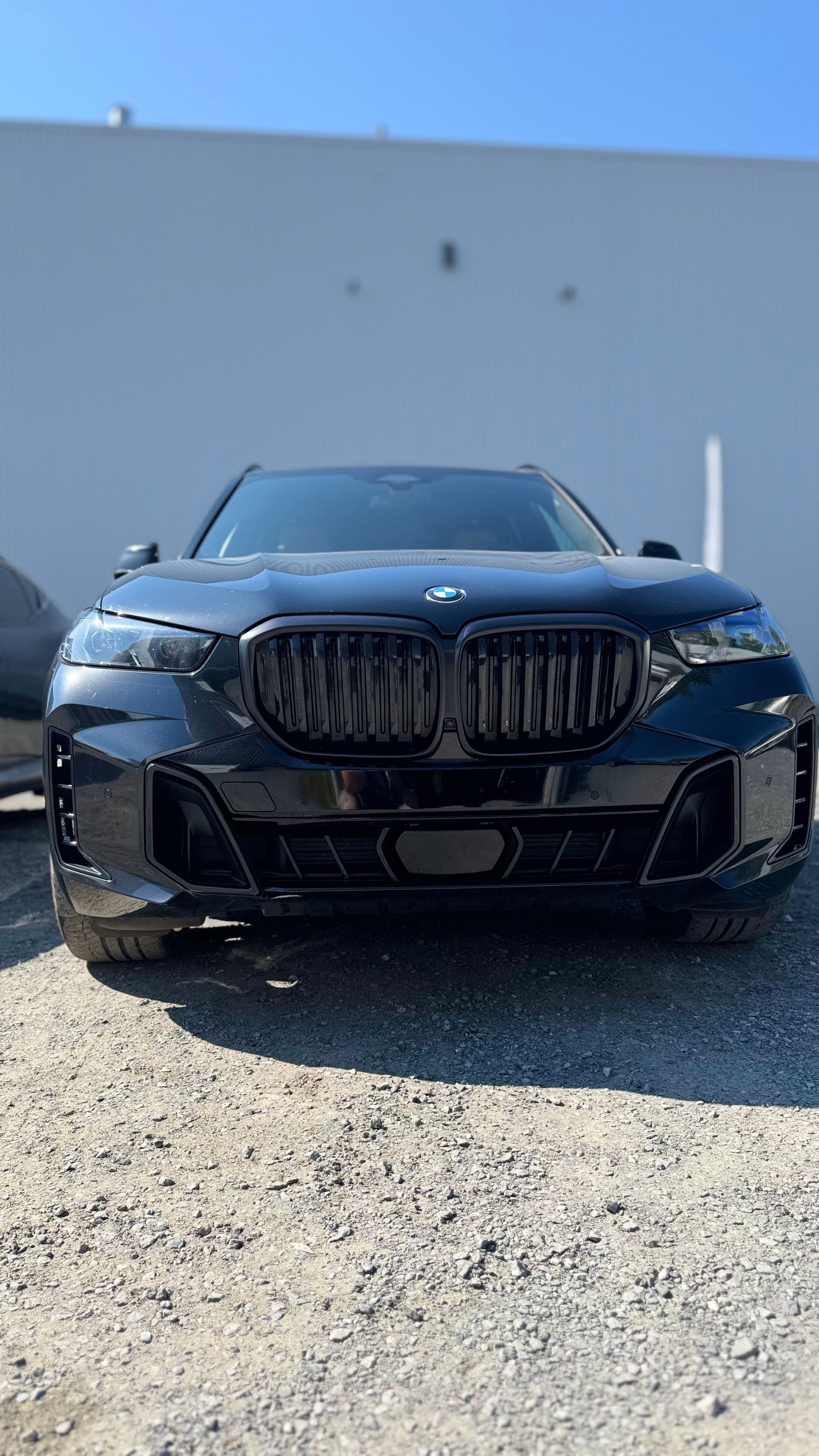 SUV BMW noir garé sur du gravier devant un bâtiment blanc, journée ensoleillée.