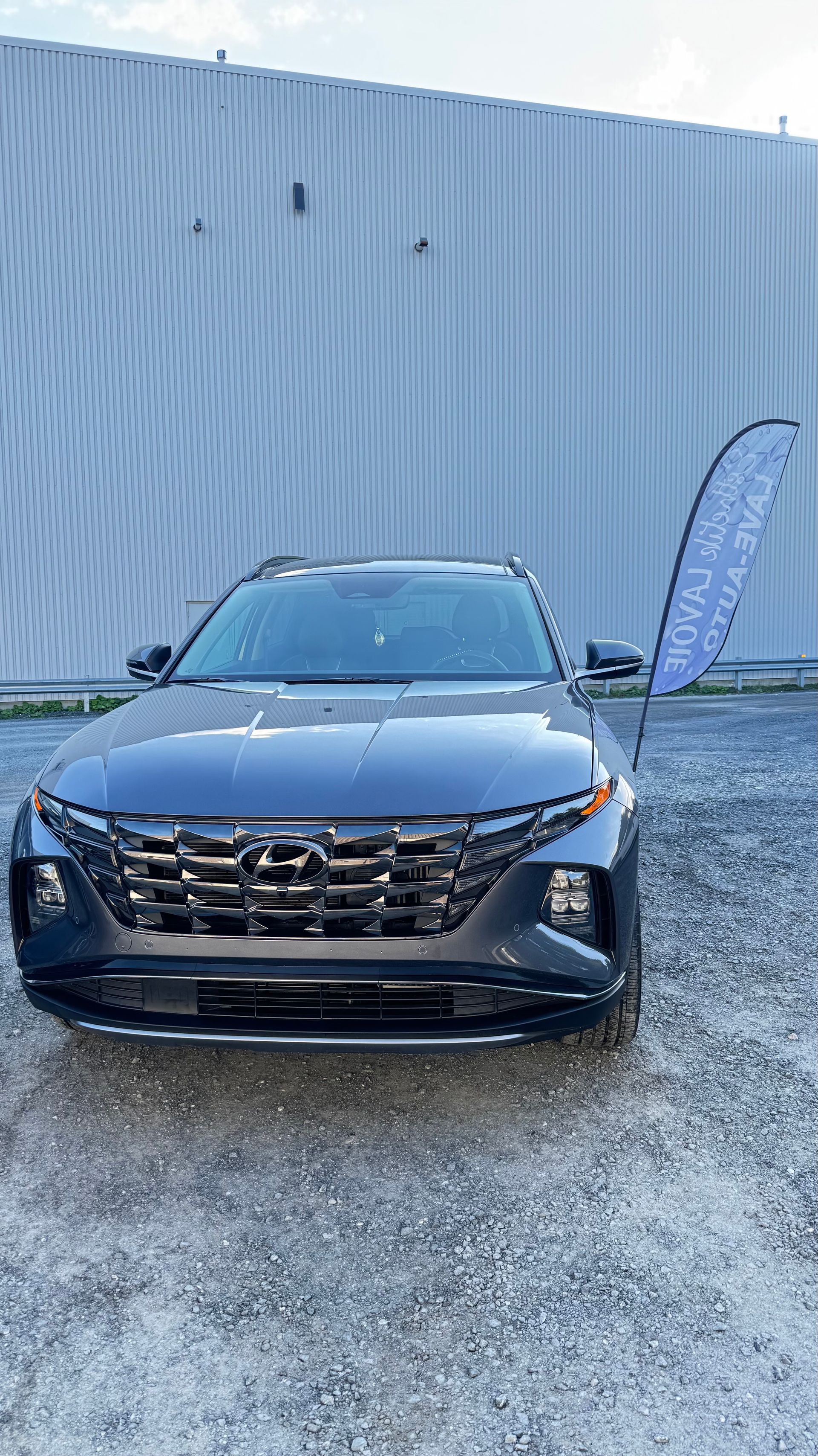 SUV Hyundai Tucson gris foncé garé devant un bâtiment gris clair, avec un grand panneau à droite.