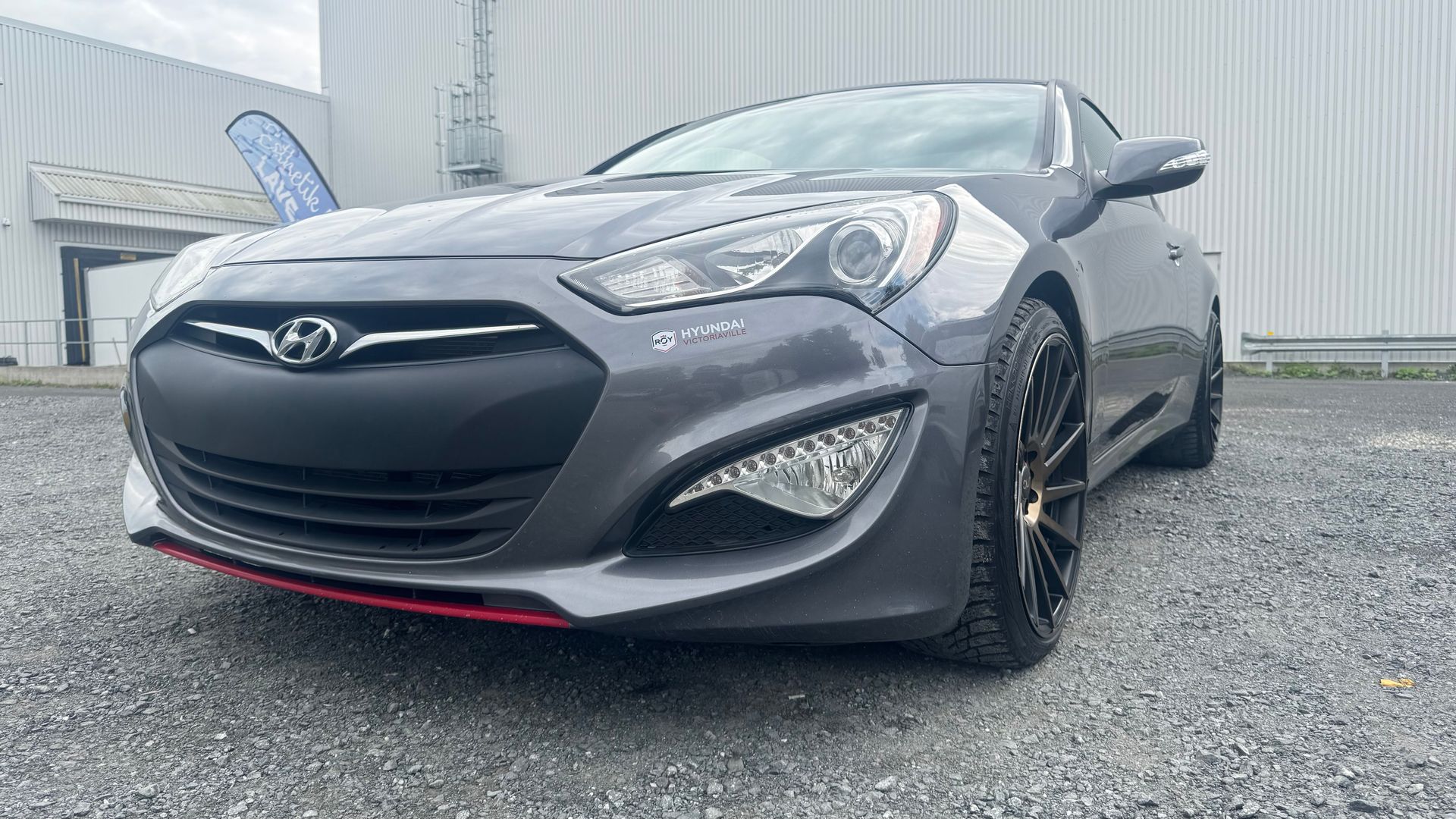 Coupé Hyundai Genesis gris avec jantes noires et garnitures rouges, garé sur du gravier.
