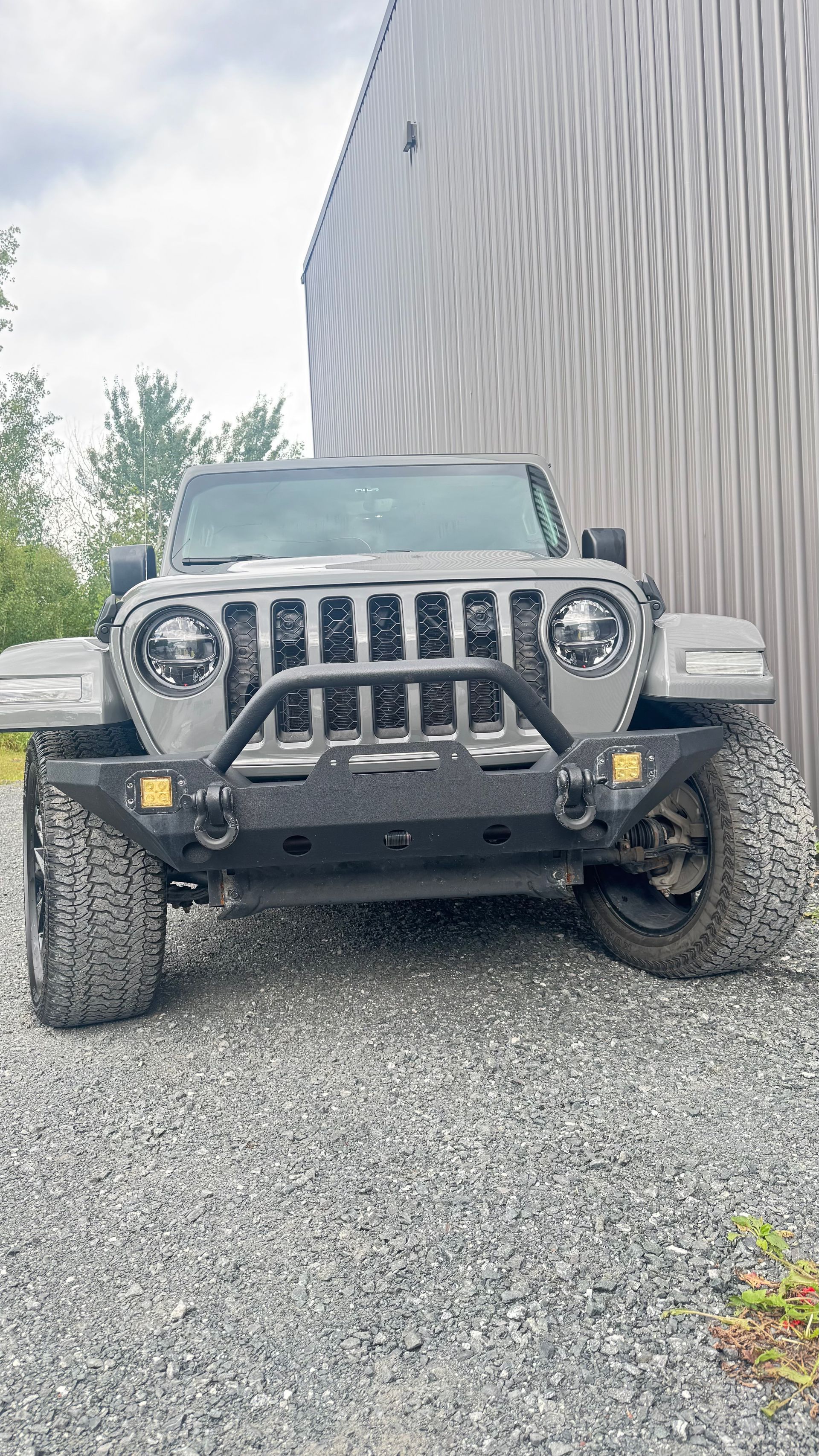 SUV Jeep gris garé sur du gravier à côté d'un bâtiment en tôle ondulée sous un ciel couvert.
