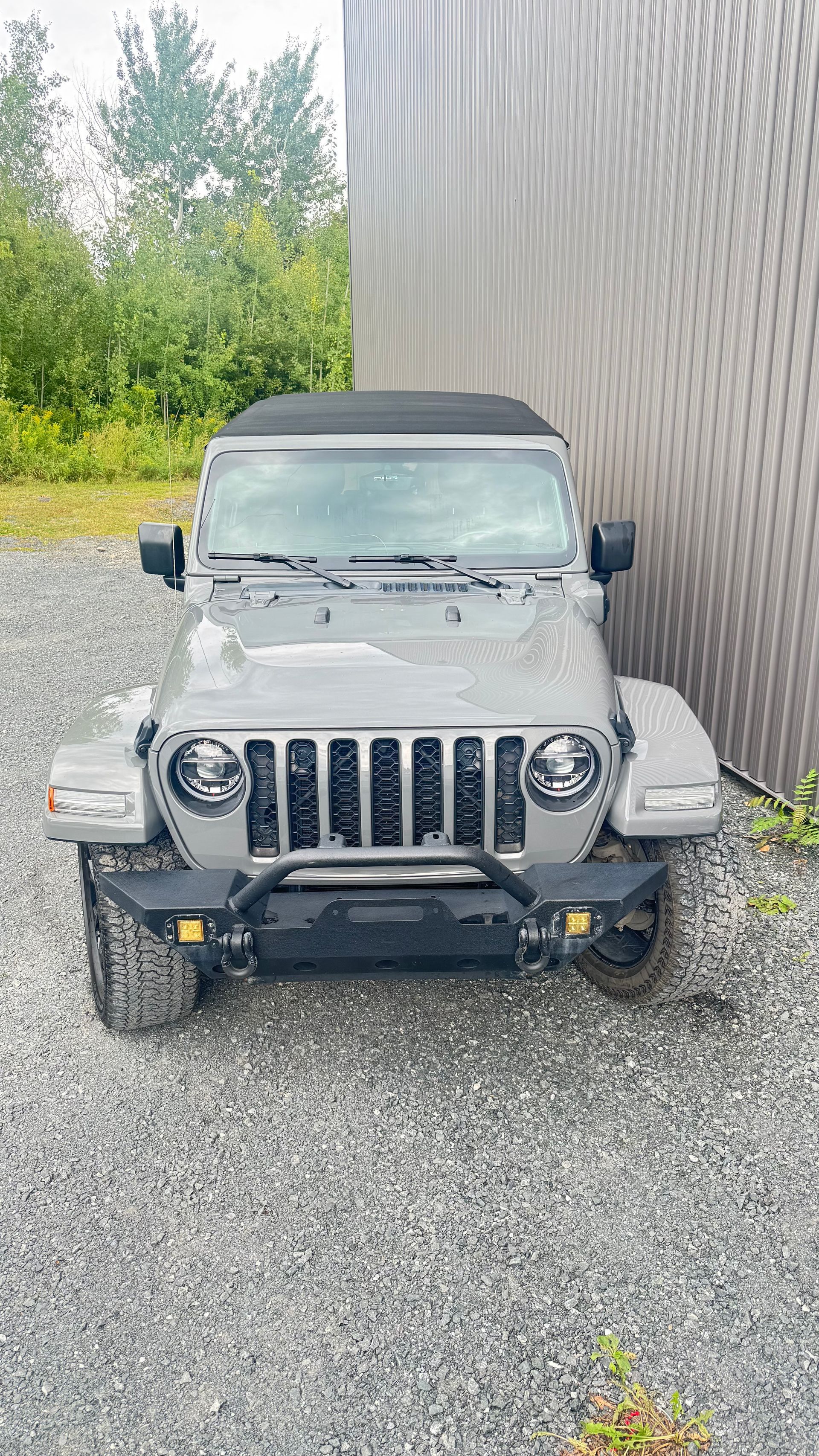 Jeep grise garée sur du gravier, près d'un mur texturé. Arbres en arrière-plan.