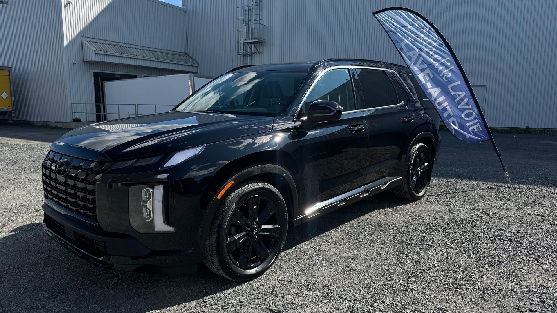 SUV Hyundai noir stationné sur du gravier devant un bâtiment avec un drapeau de concessionnaire.